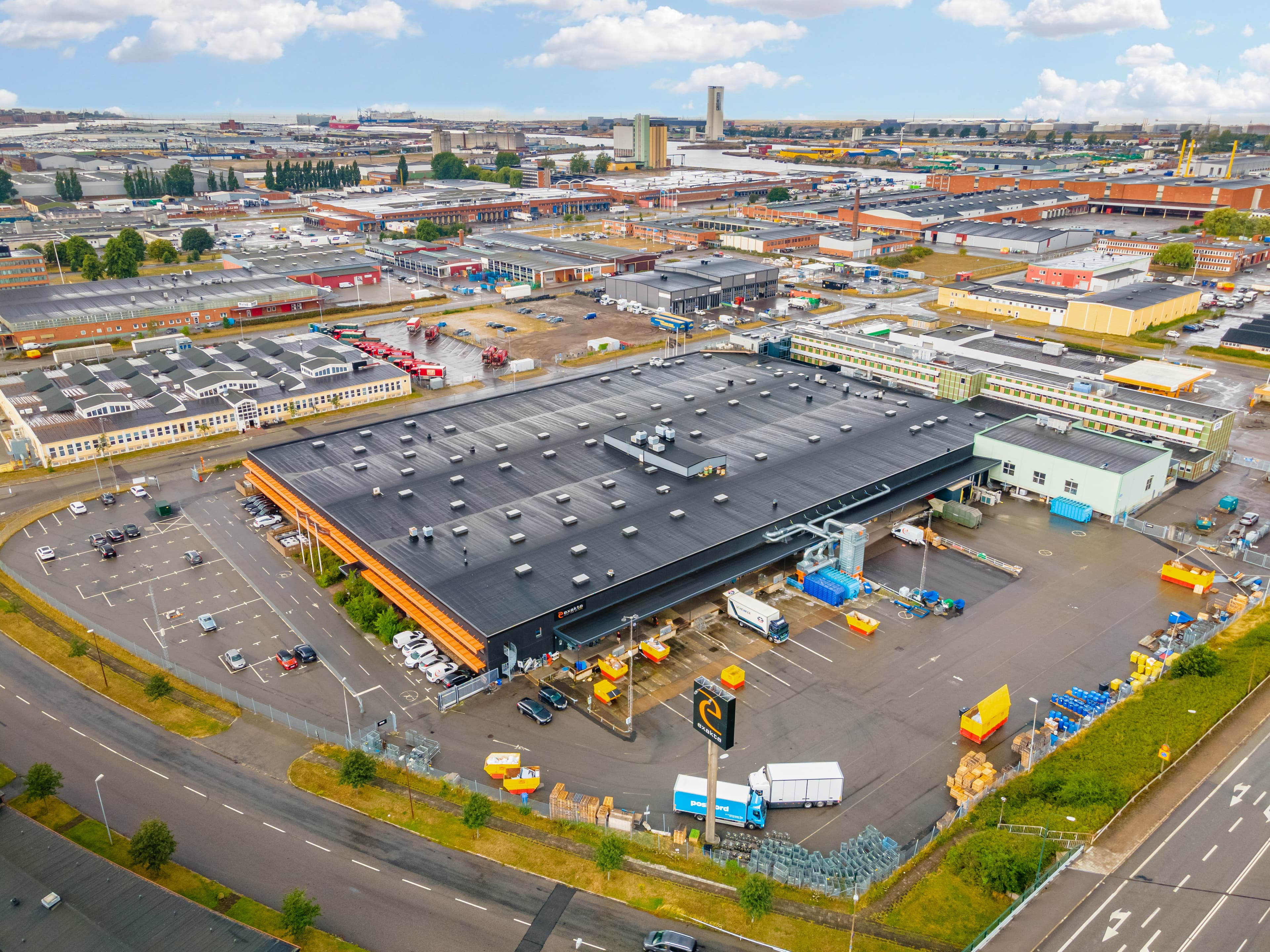Aerial view of an industrial area with a large warehouse, parking lots, and surrounding roads under a partly cloudy sky.