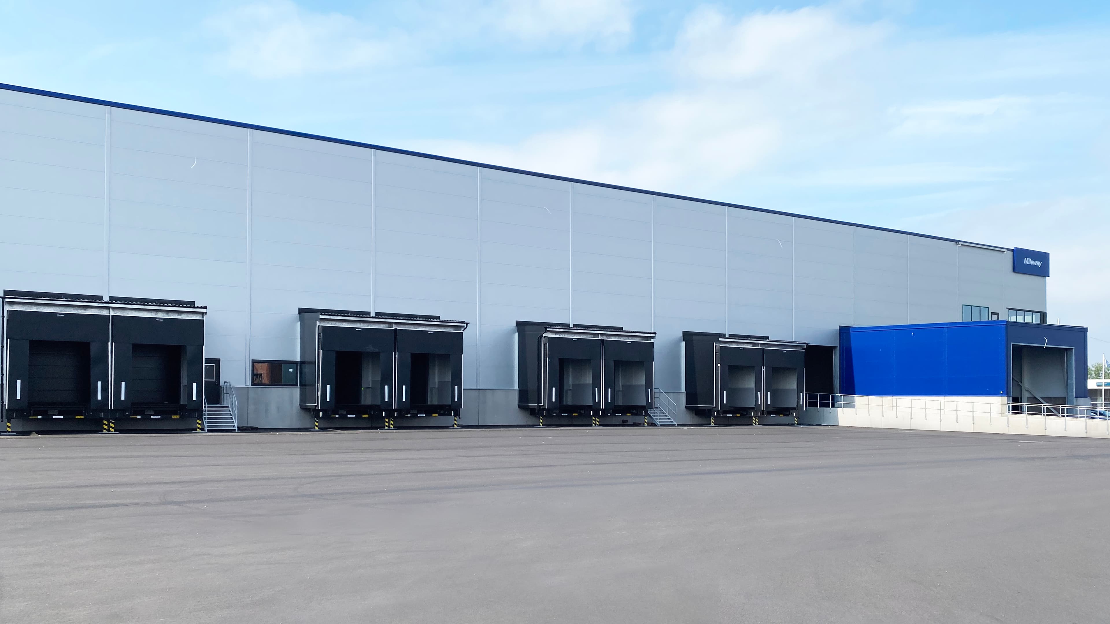 A large industrial warehouse with five loading bays is shown, featuring a blue section at one end under a clear sky.