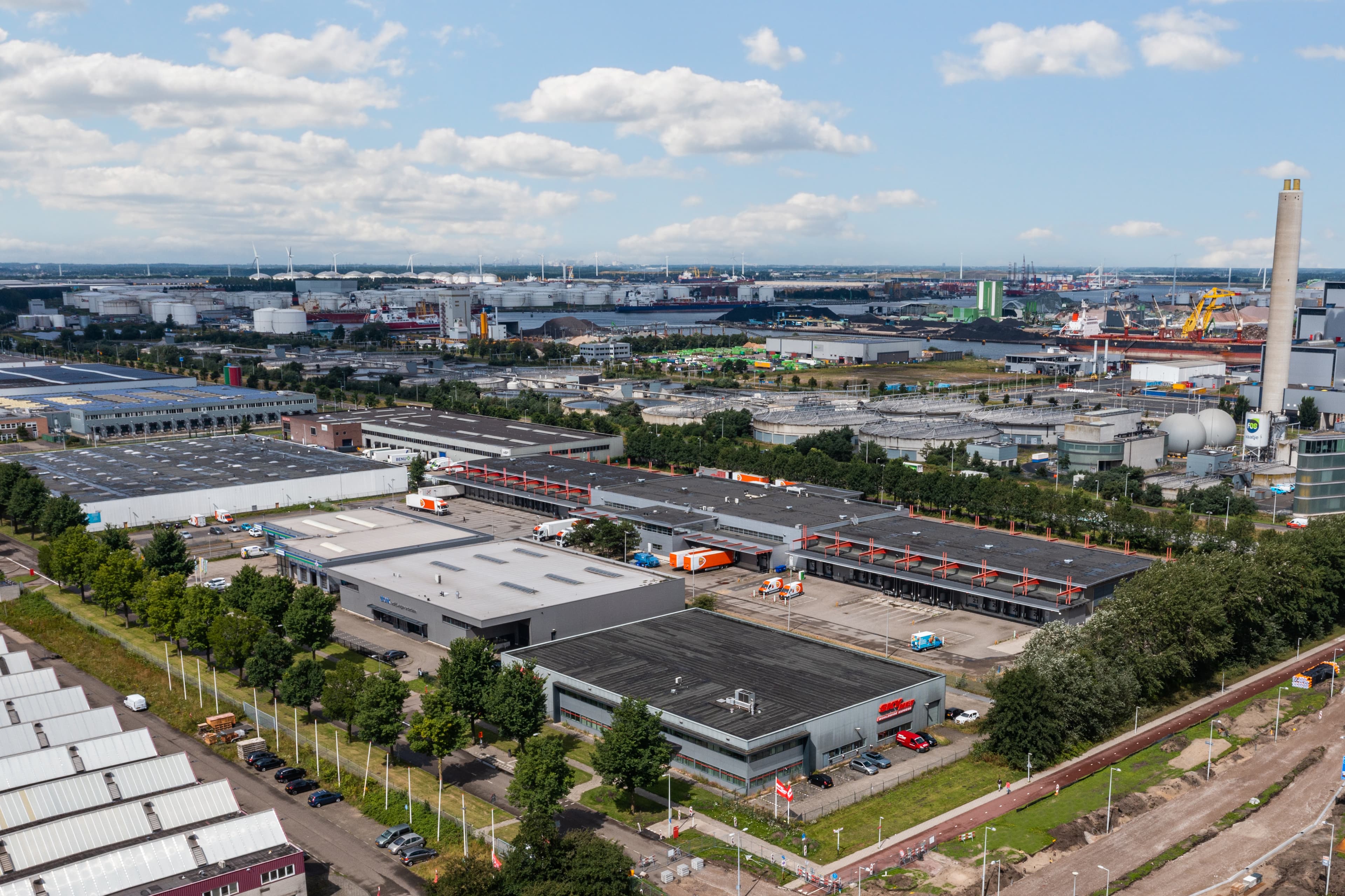 Vista aérea de un complejo industrial con almacenes, zonas verdes y vehículos estacionados bajo un cielo parcialmente nublado.