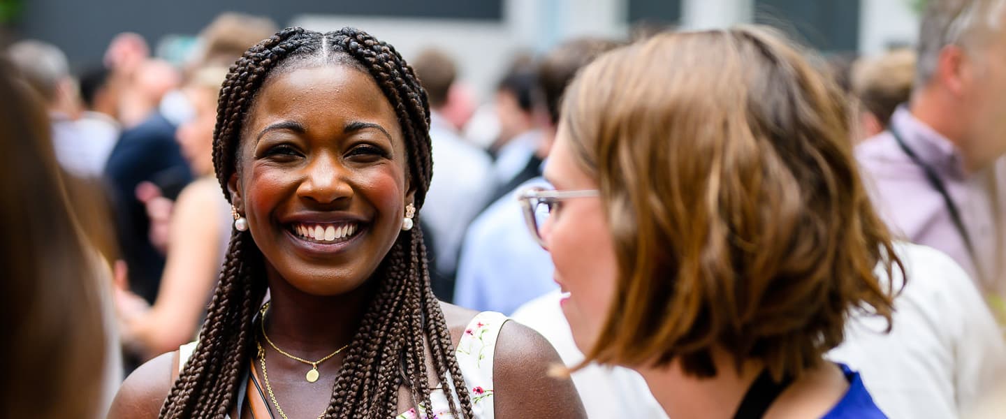 A smiling person with braided hair stands in a crowd, wearing a white top with floral patterns and a necklace, engaged in conversation with someone wearing glasses.