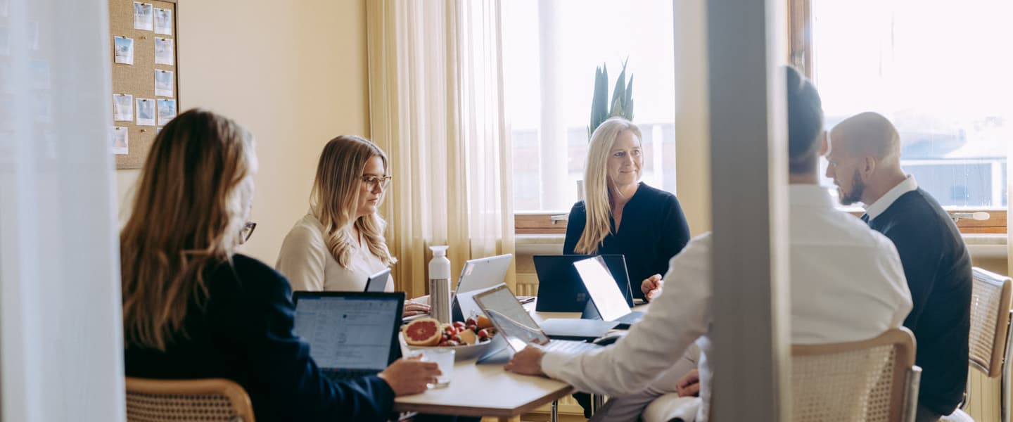 Five people having a meeting in an office, using laptops and tablets, with a plate of fruit on the table. Natural light comes through a large window.