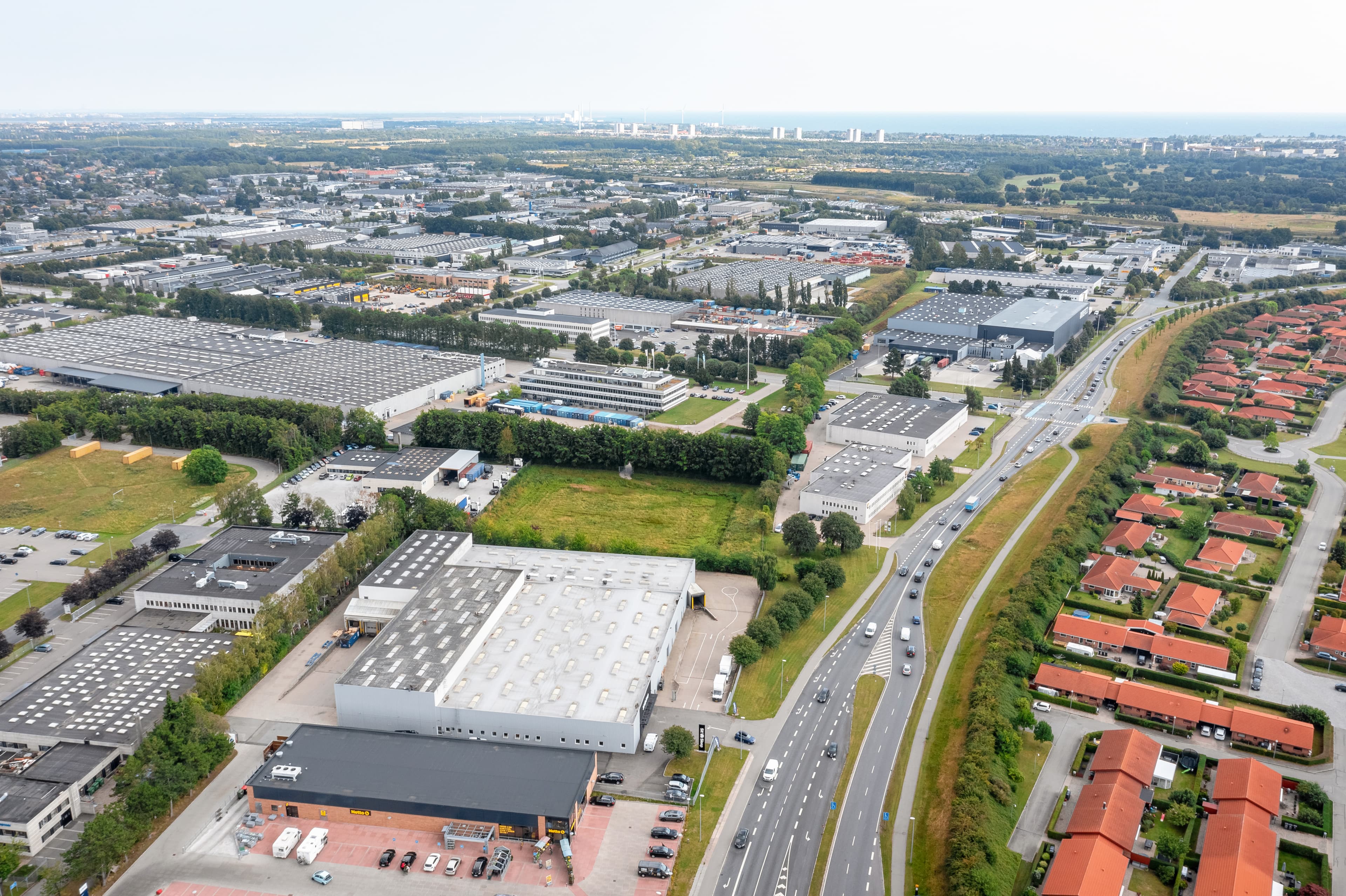 Vista aérea de un área industrial con almacenes, carreteras y edificios residenciales cercanos, rodeados de árboles y espacios abiertos.