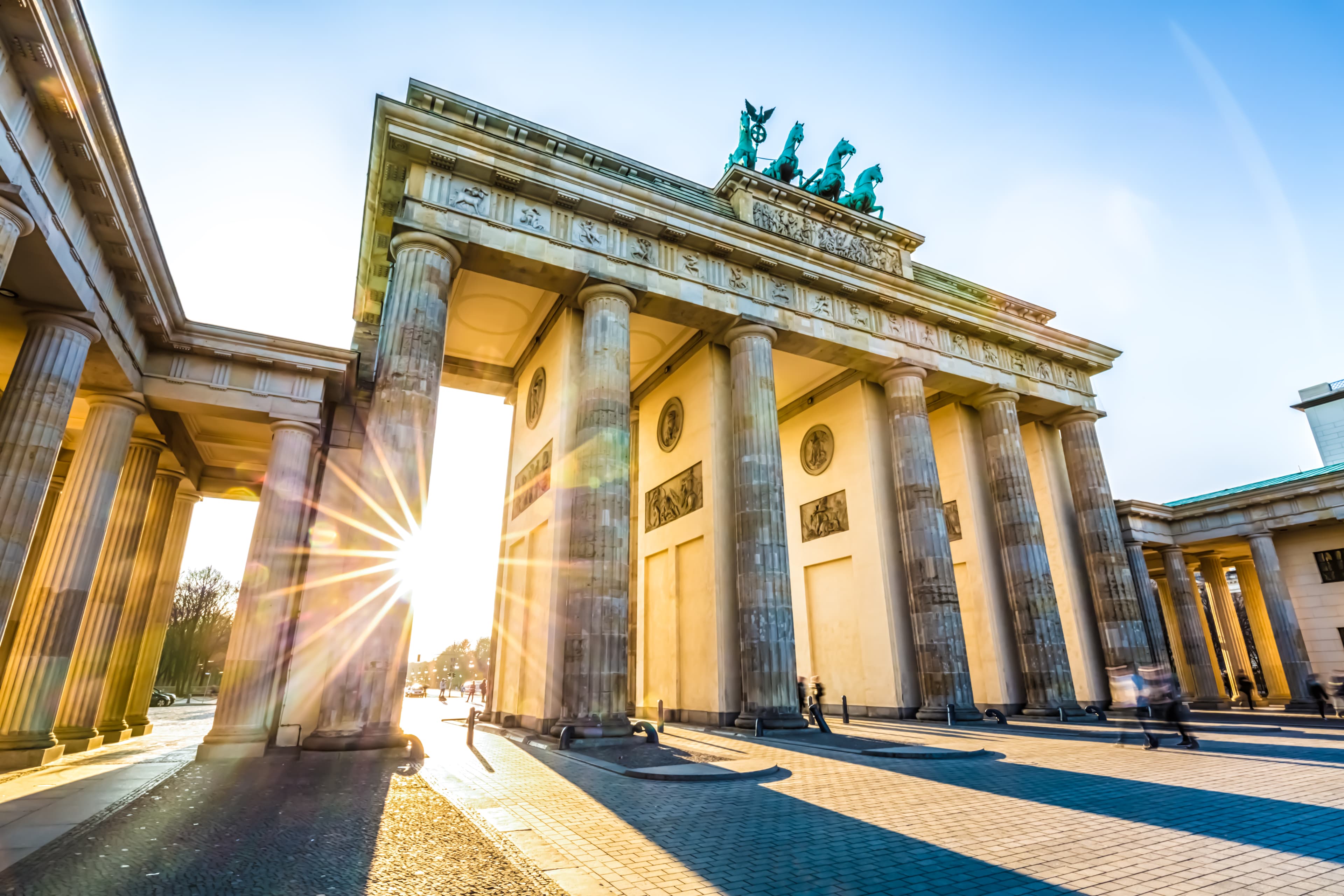 The image shows the Brandenburg Gate with a sunburst visible through its columns against a clear blue sky.