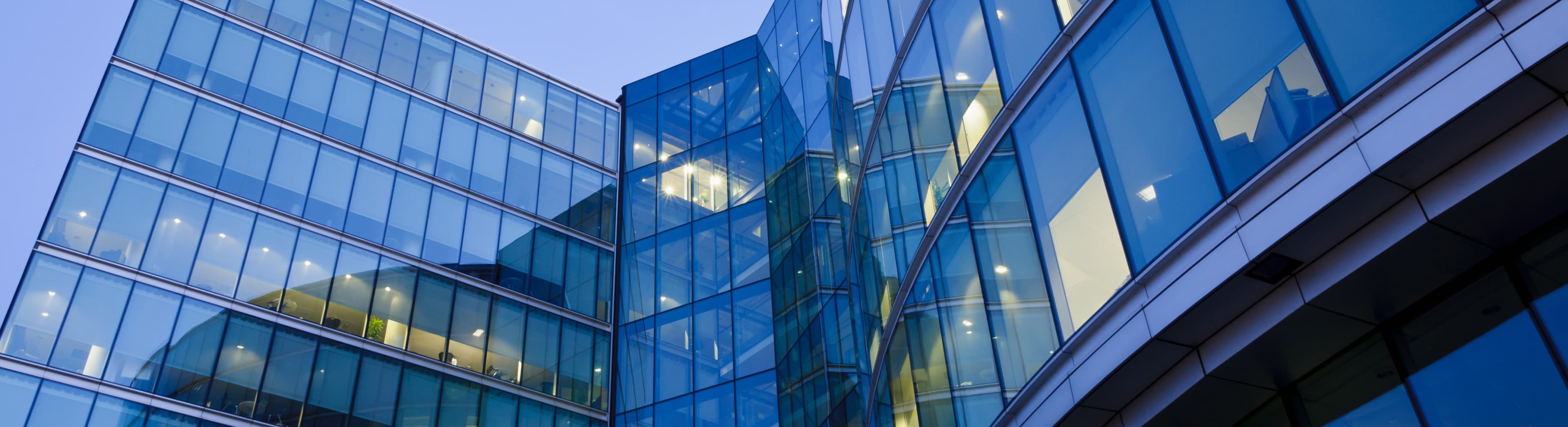 A modern glass office building with reflective windows, captured from a low angle, set against a clear sky.