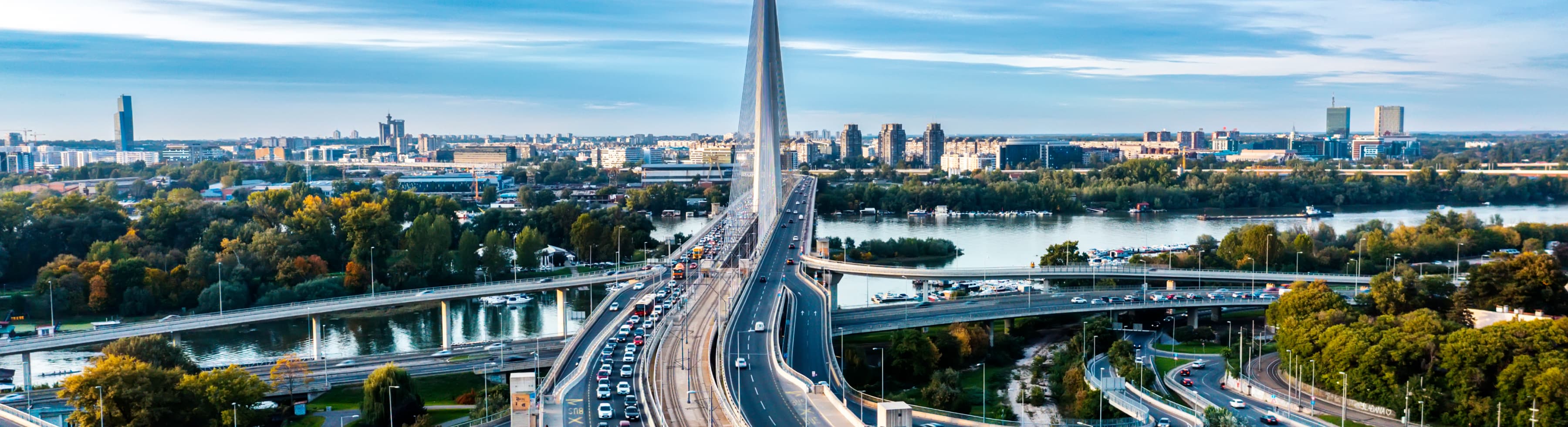 Aerial view of a large cable-stayed bridge over a river, surrounded by a cityscape with tall buildings and greenery. Cars are visible on the bridge and roads below.