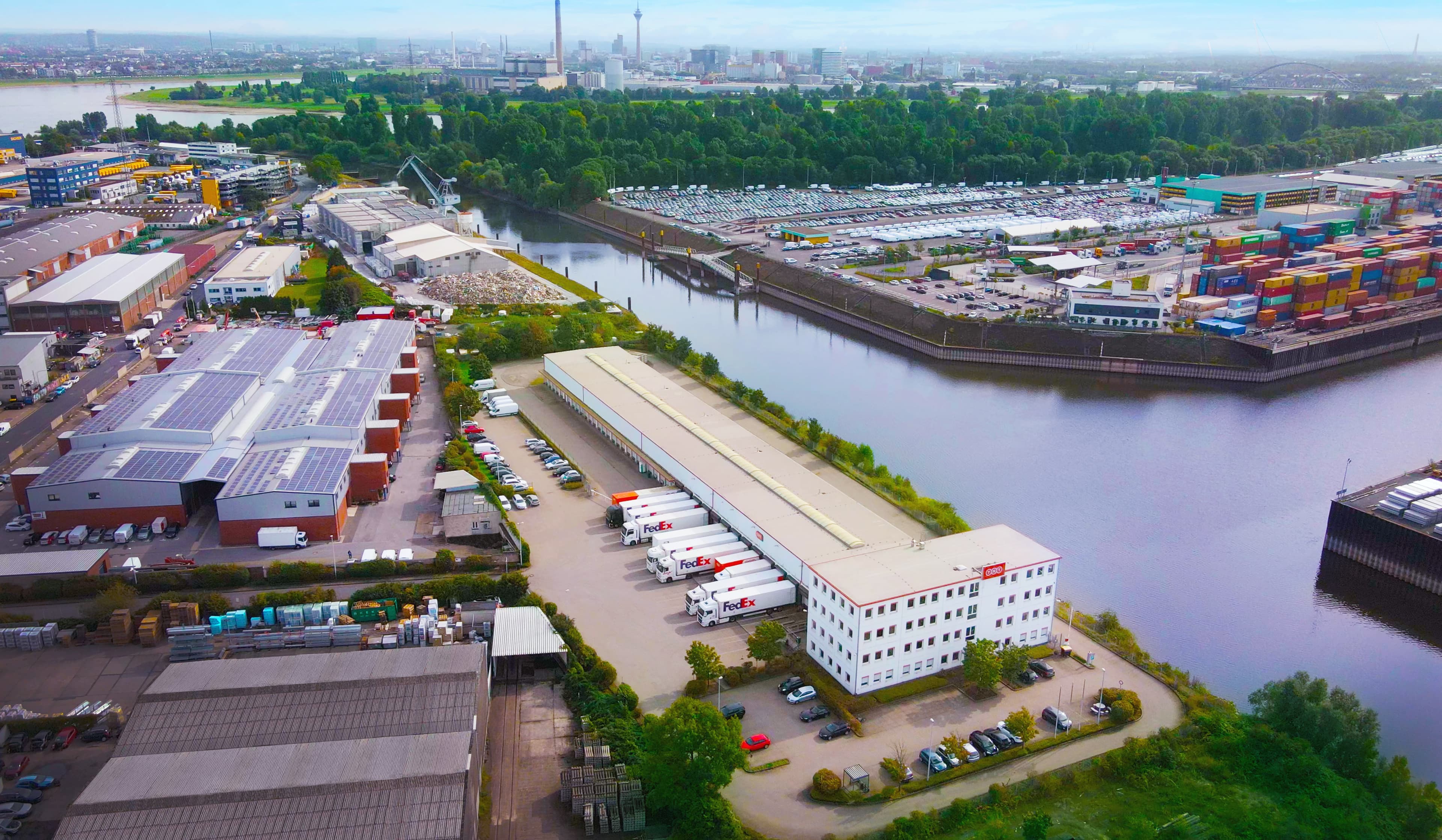 Vista aérea de una zona industrial con almacenes, camiones y contenedores de carga cerca de un río. Se ve el horizonte de la ciudad al fondo.