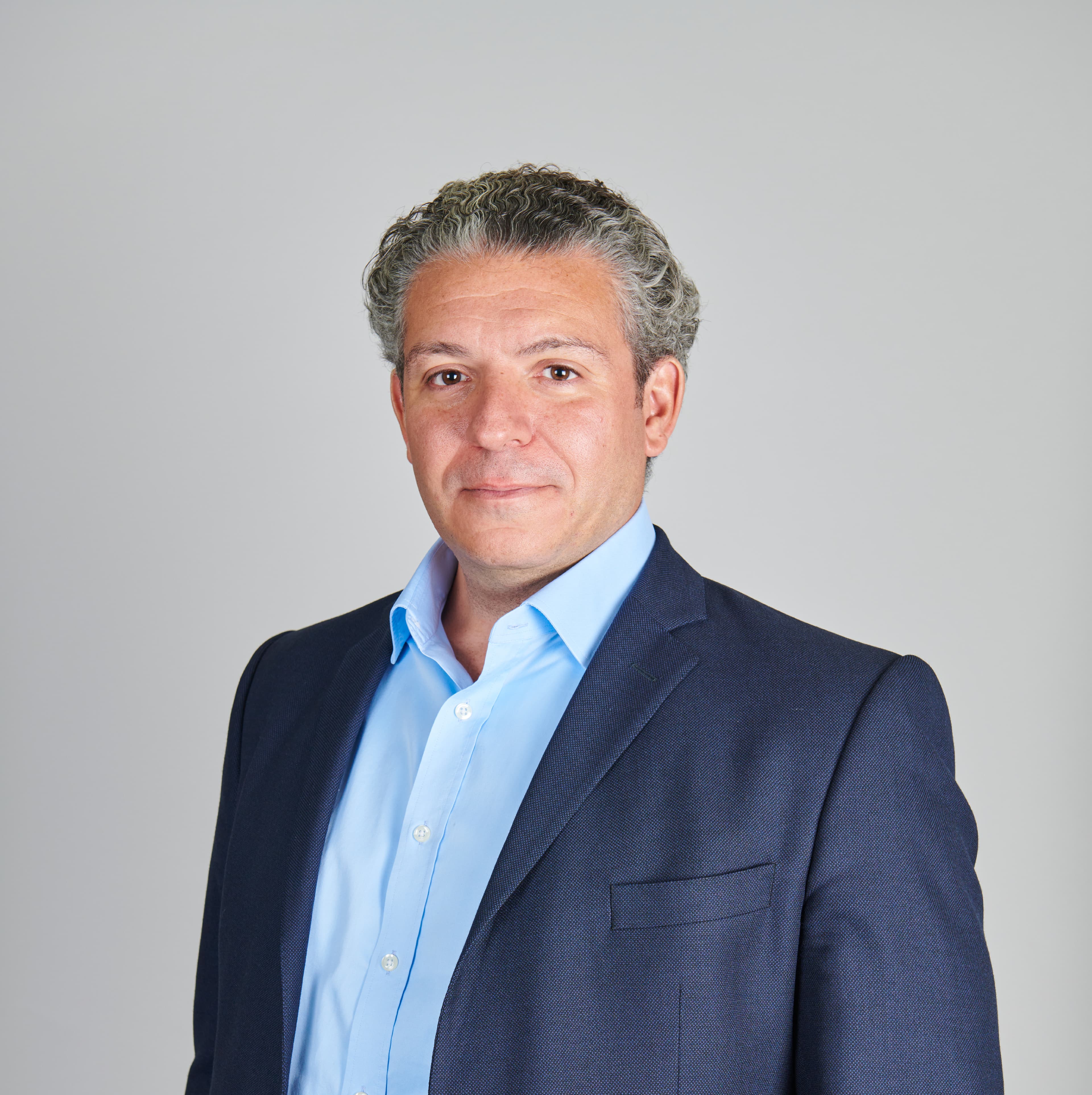 A man in a blue shirt and dark blazer stands against a plain gray background.