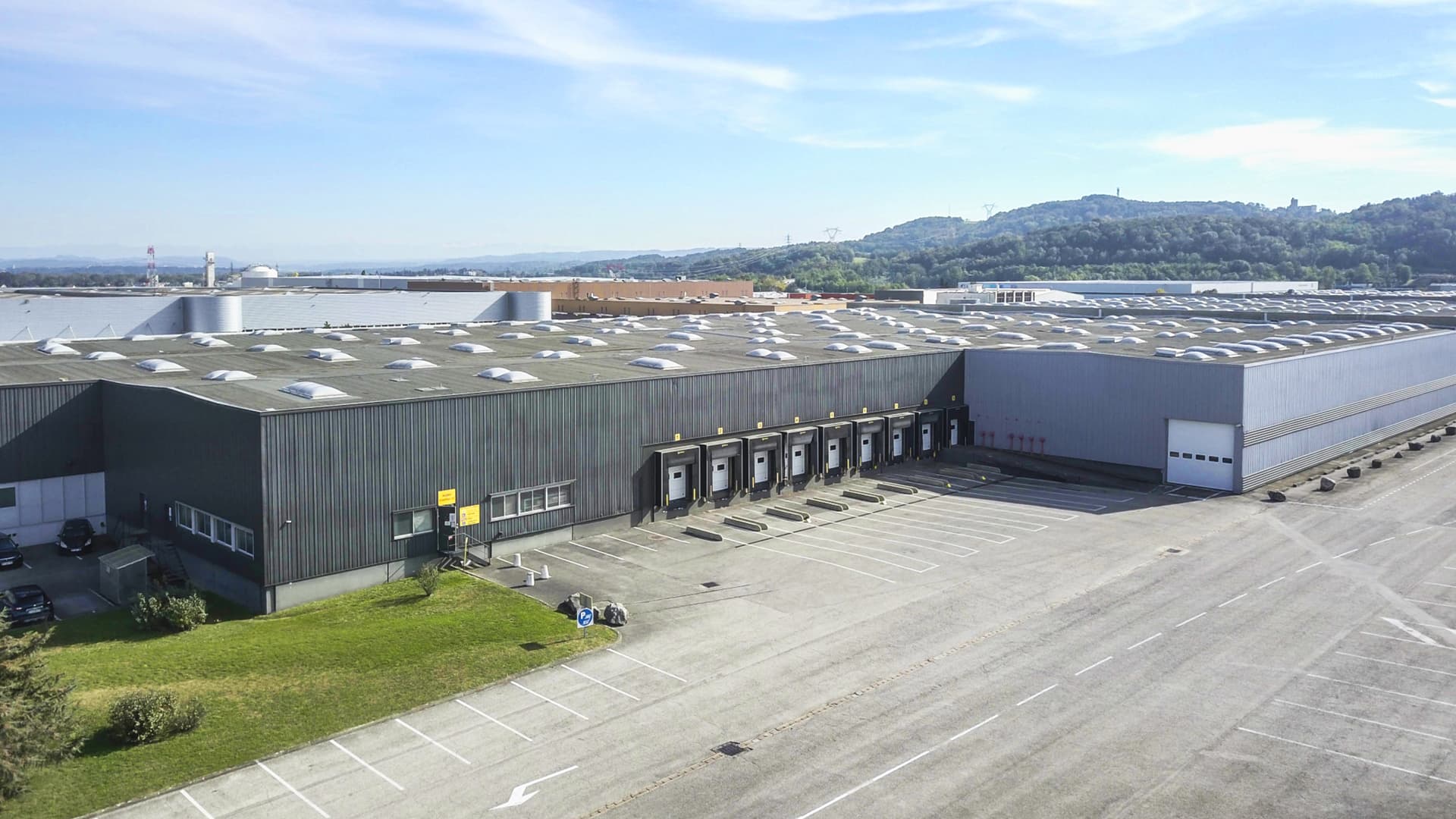 Aerial view of a large industrial warehouse with multiple loading docks, set in a spacious parking area, surrounded by hills under a clear blue sky.
