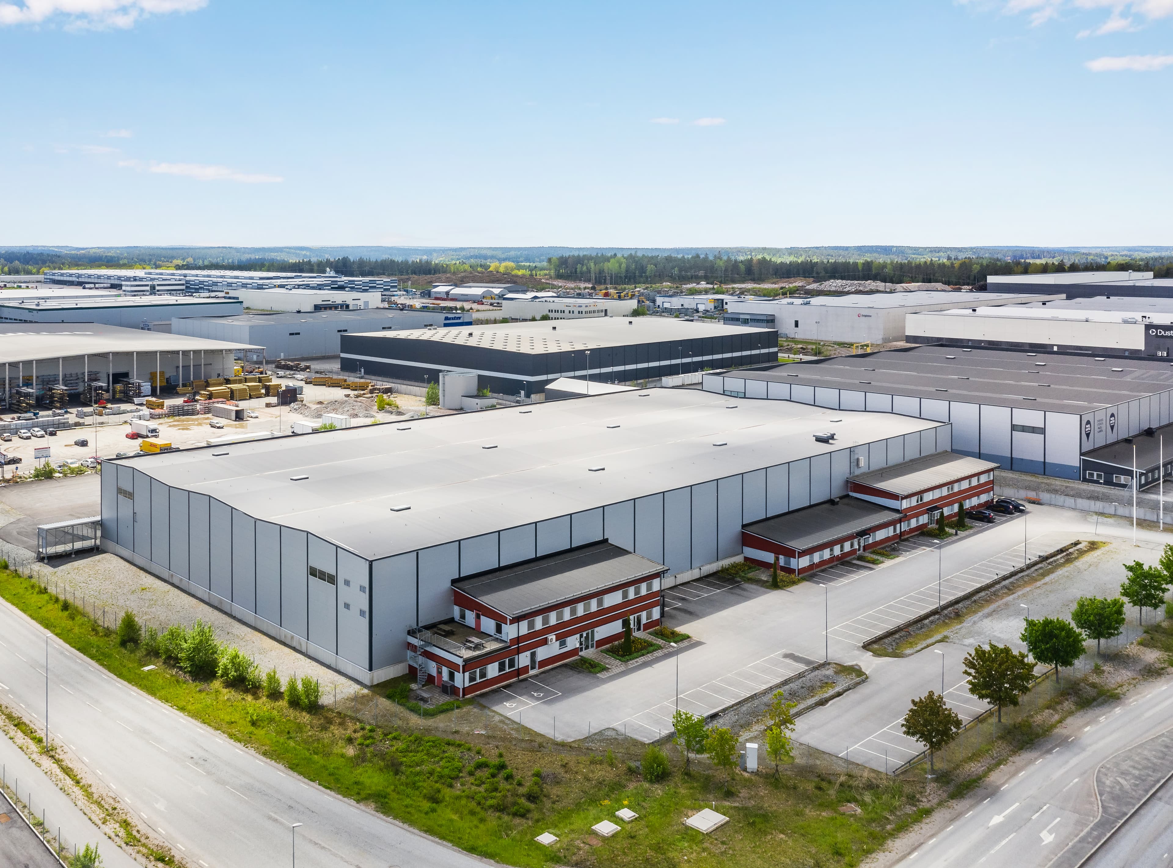 Aerial view of a large industrial complex with multiple warehouse buildings and parking areas, surrounded by roads and greenery under a clear blue sky.