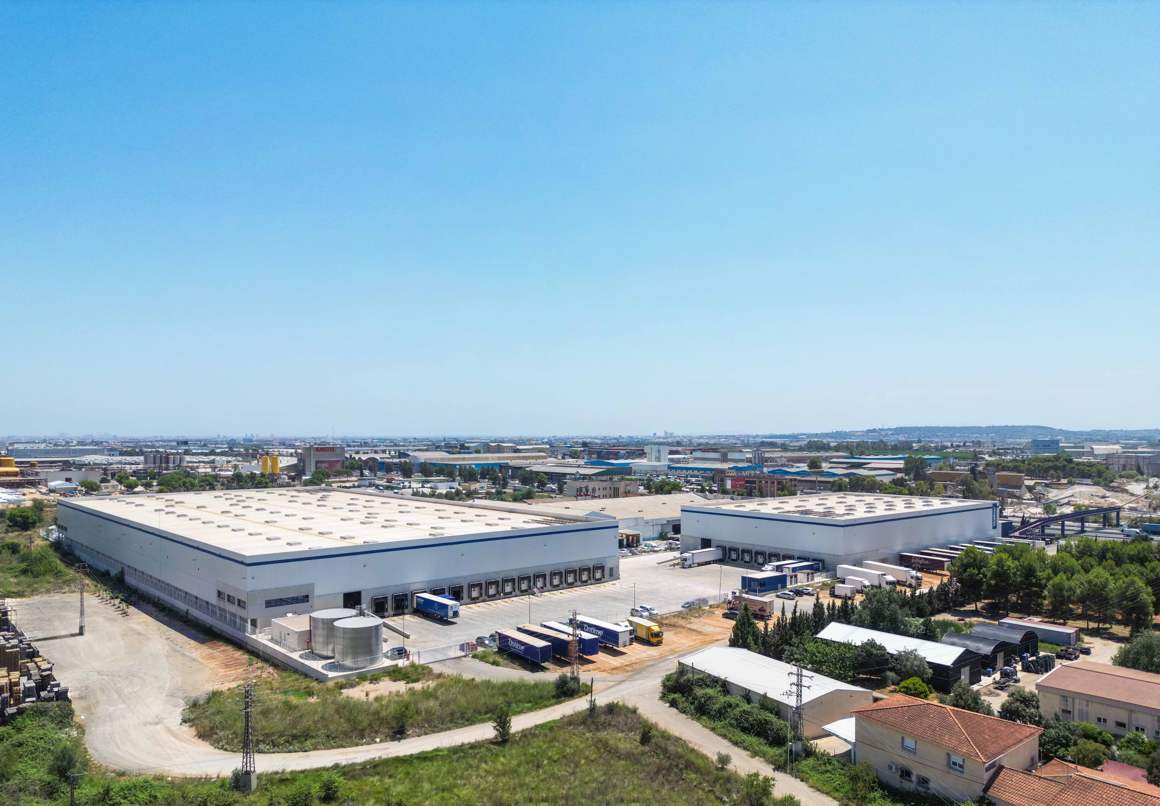 Aerial view of an industrial area featuring two large warehouses surrounded by trucks, greenery, and smaller buildings under a clear blue sky.