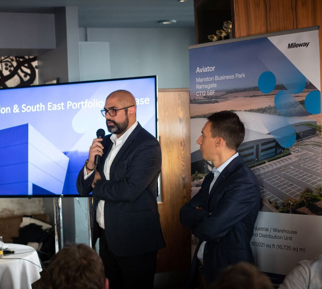 Two men in suits present at an event. One speaks into a microphone, while the other listens. A screen and a banner with building images are in the background.