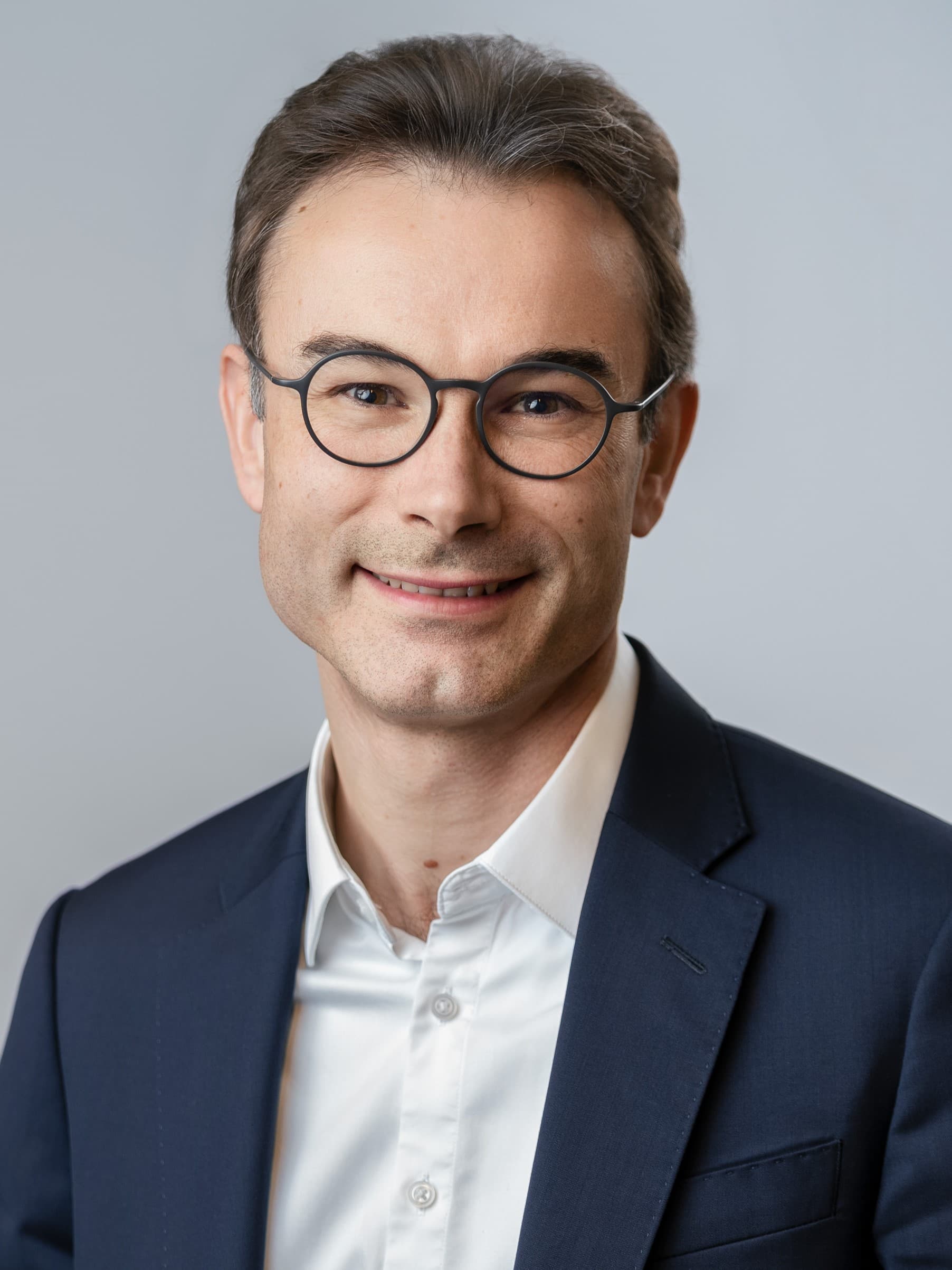 A person wearing glasses, a dark suit, and a white shirt smiles at the camera against a plain light background.