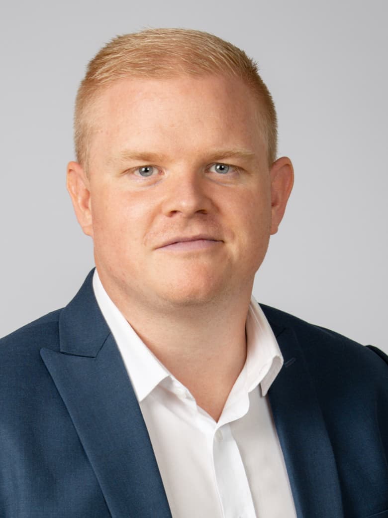 A person with short blond hair in a blue suit and white shirt against a neutral background.