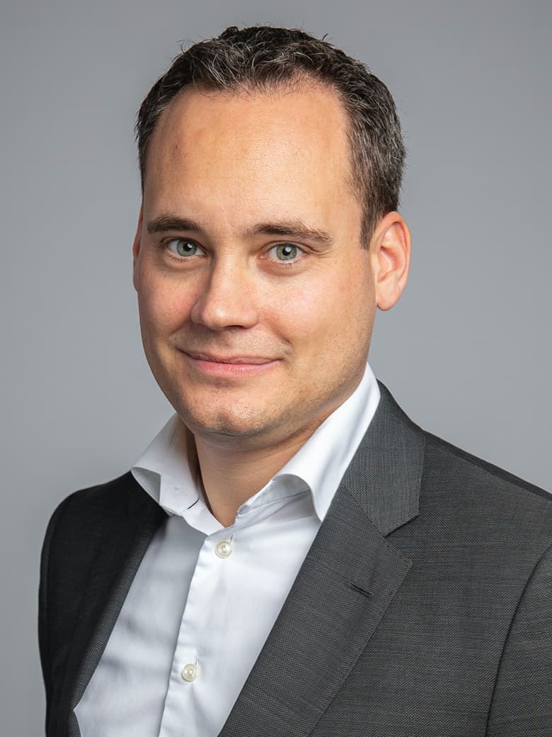 A person in a dark suit and white shirt poses against a gray background.