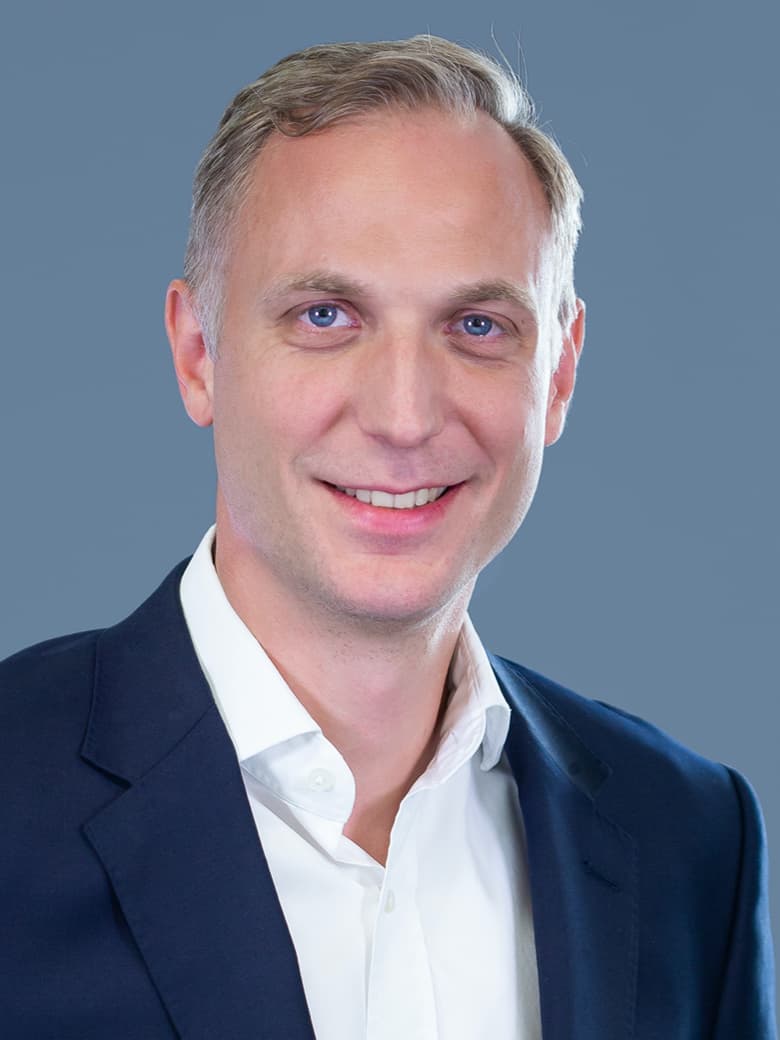 A man in a suit and white shirt smiling against a plain blue background.