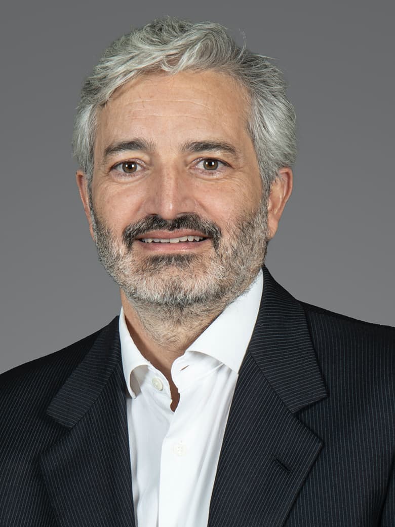 A man with gray hair and a beard is wearing a dark suit jacket and a white shirt. The background is plain gray.
