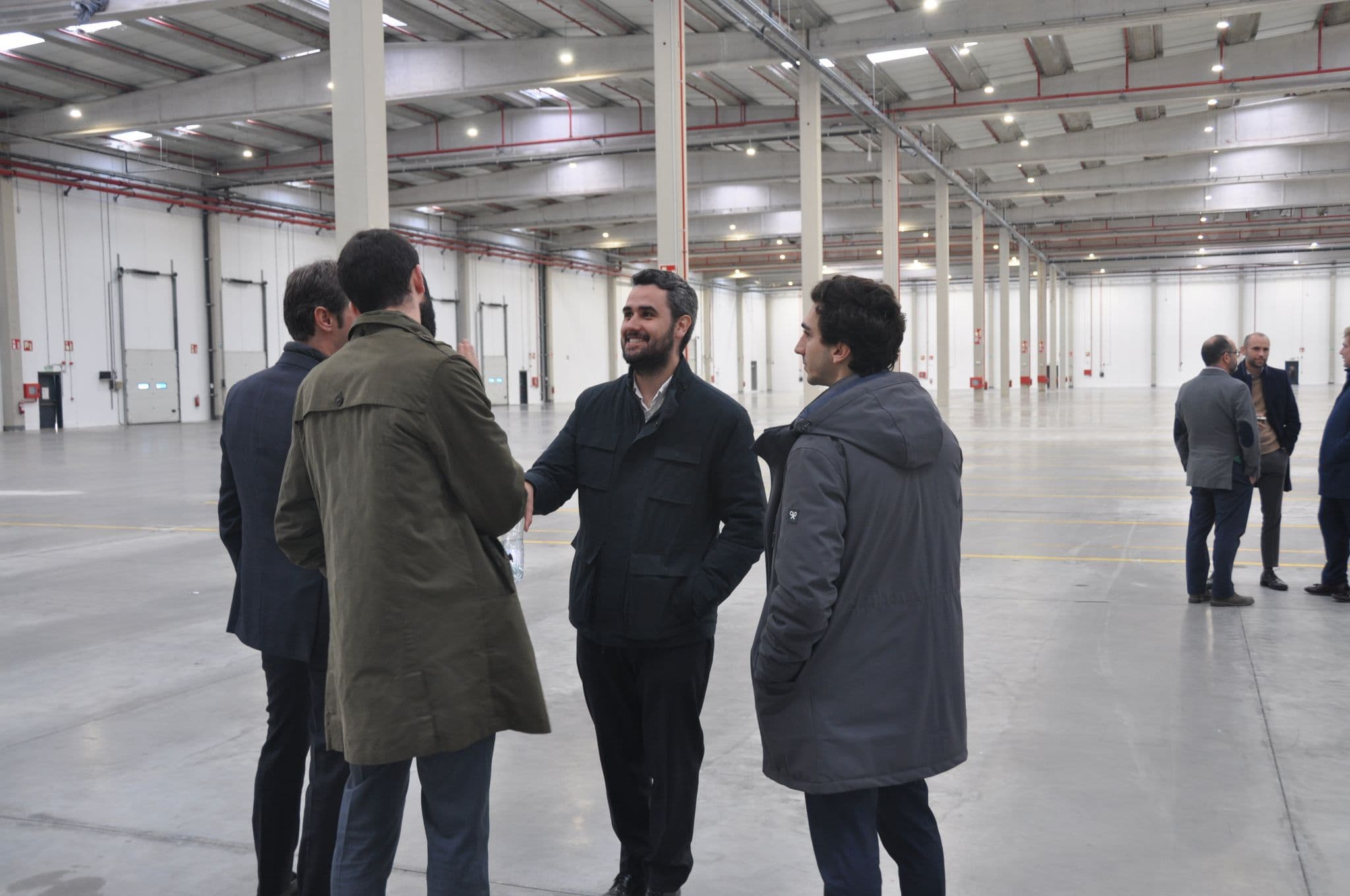 A group of men stands and talks in a large, empty warehouse with high ceilings and bright lighting.