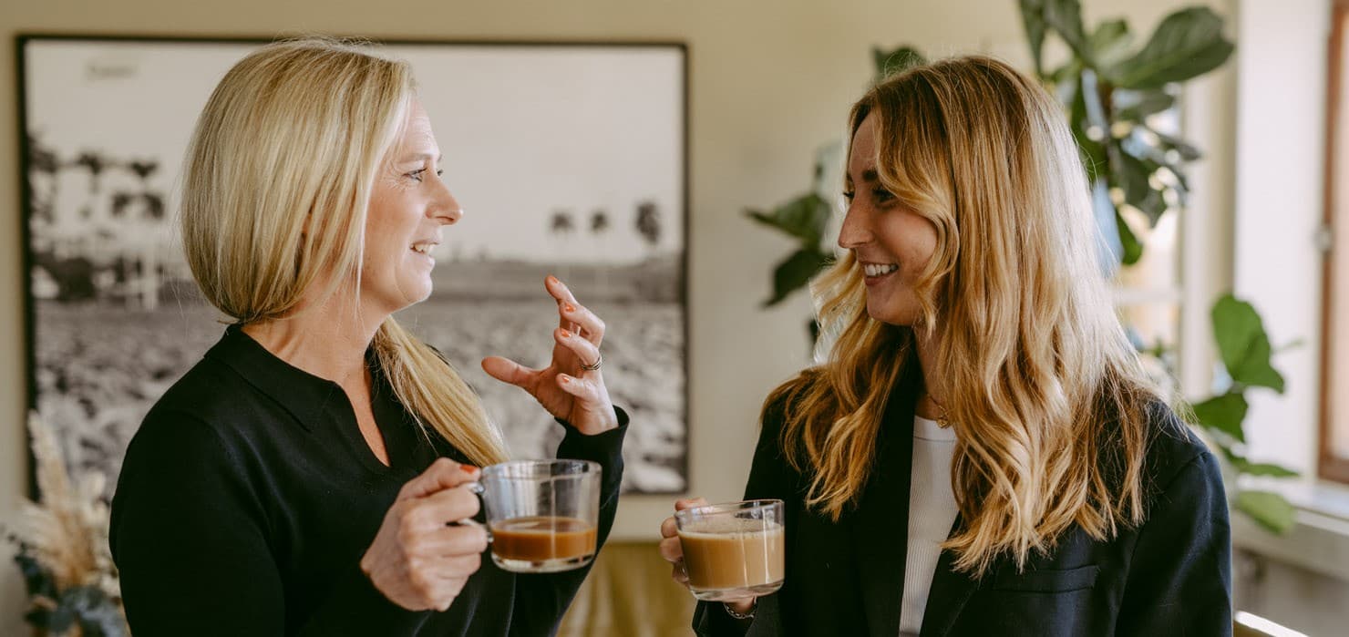 Two women holding coffee mugs engage in conversation, standing indoors with a large framed photograph and plants in the background.