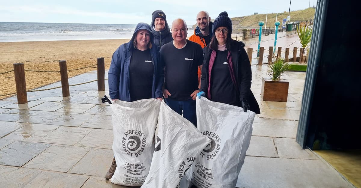 A group of five people standing on a wet beachfront holding litter bags.