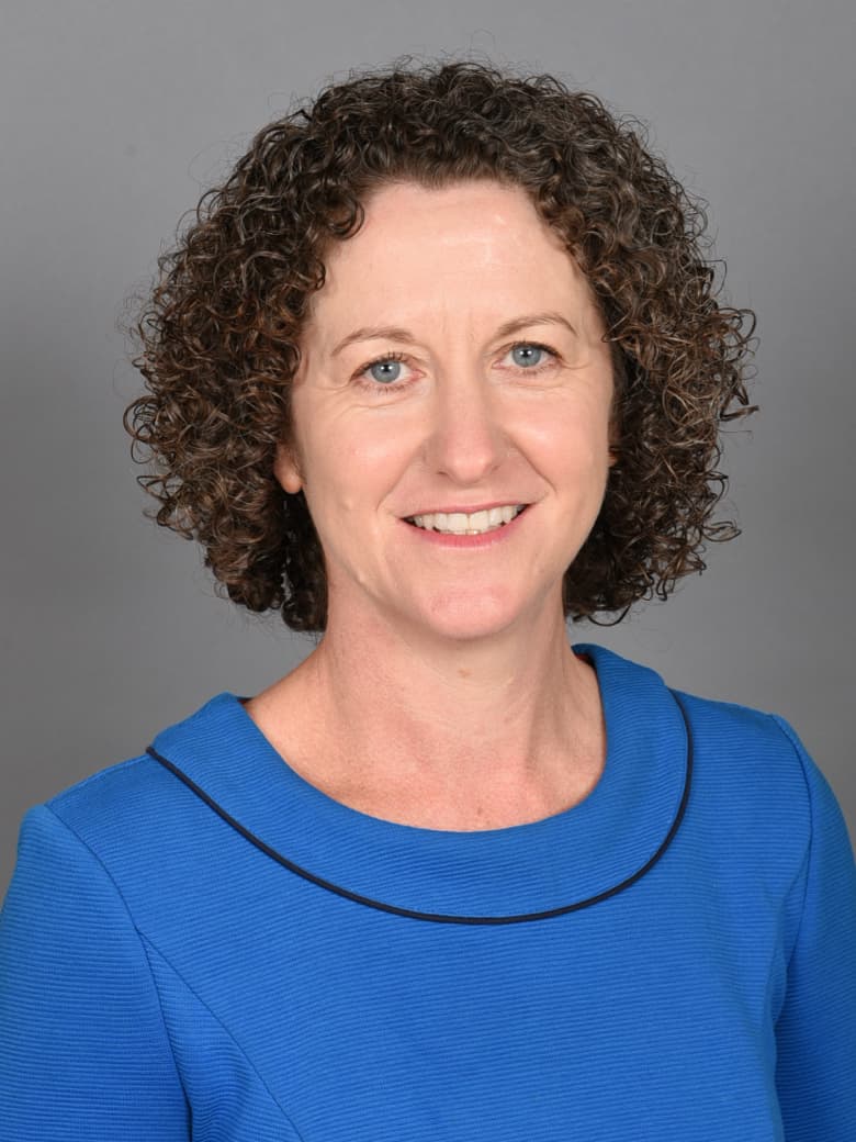 A person with curly hair, wearing a blue top, smiles against a gray background.