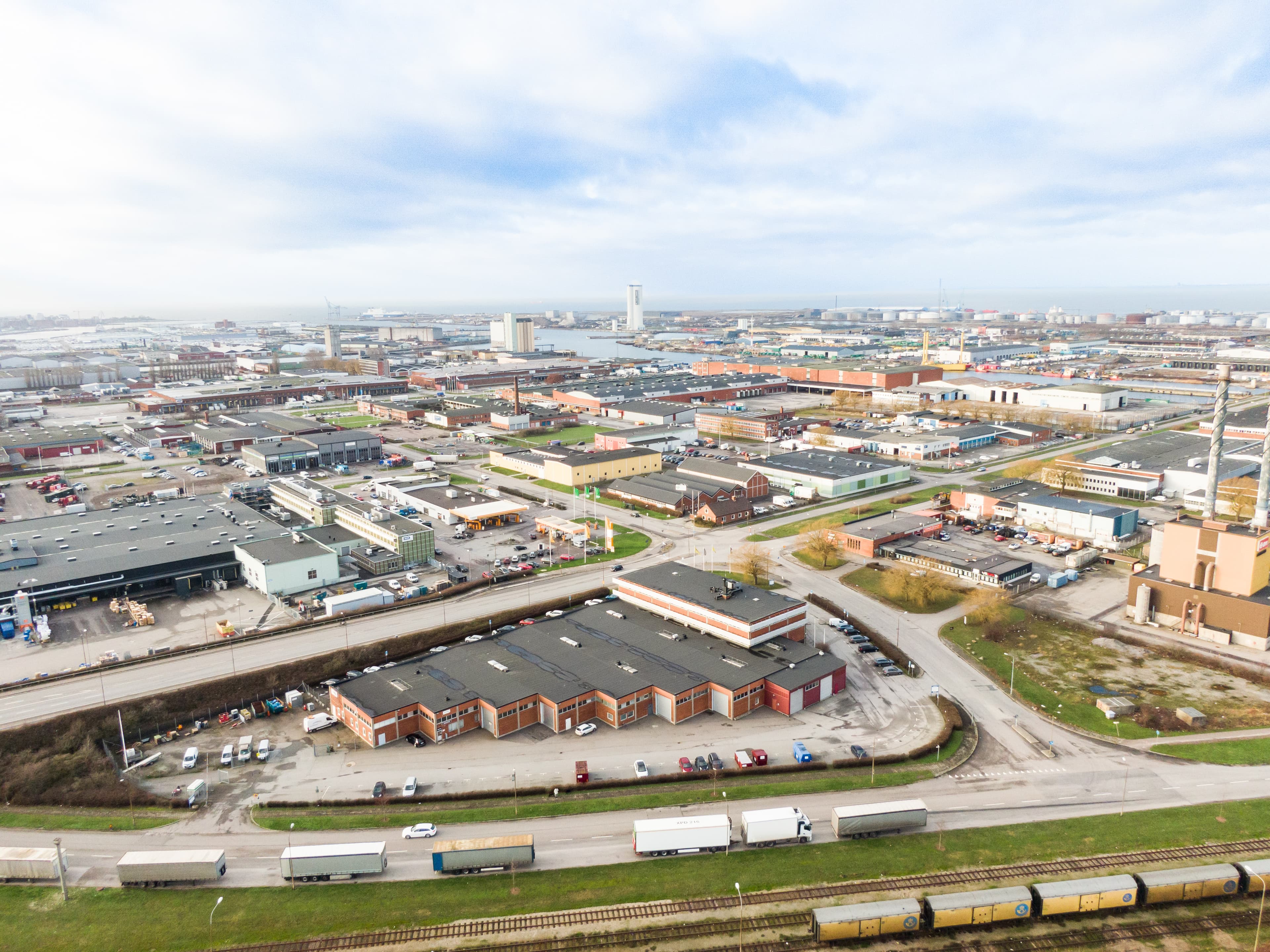 Aerial view of an industrial area with warehouses, roads, parked trucks, and railway tracks under a cloudy sky.