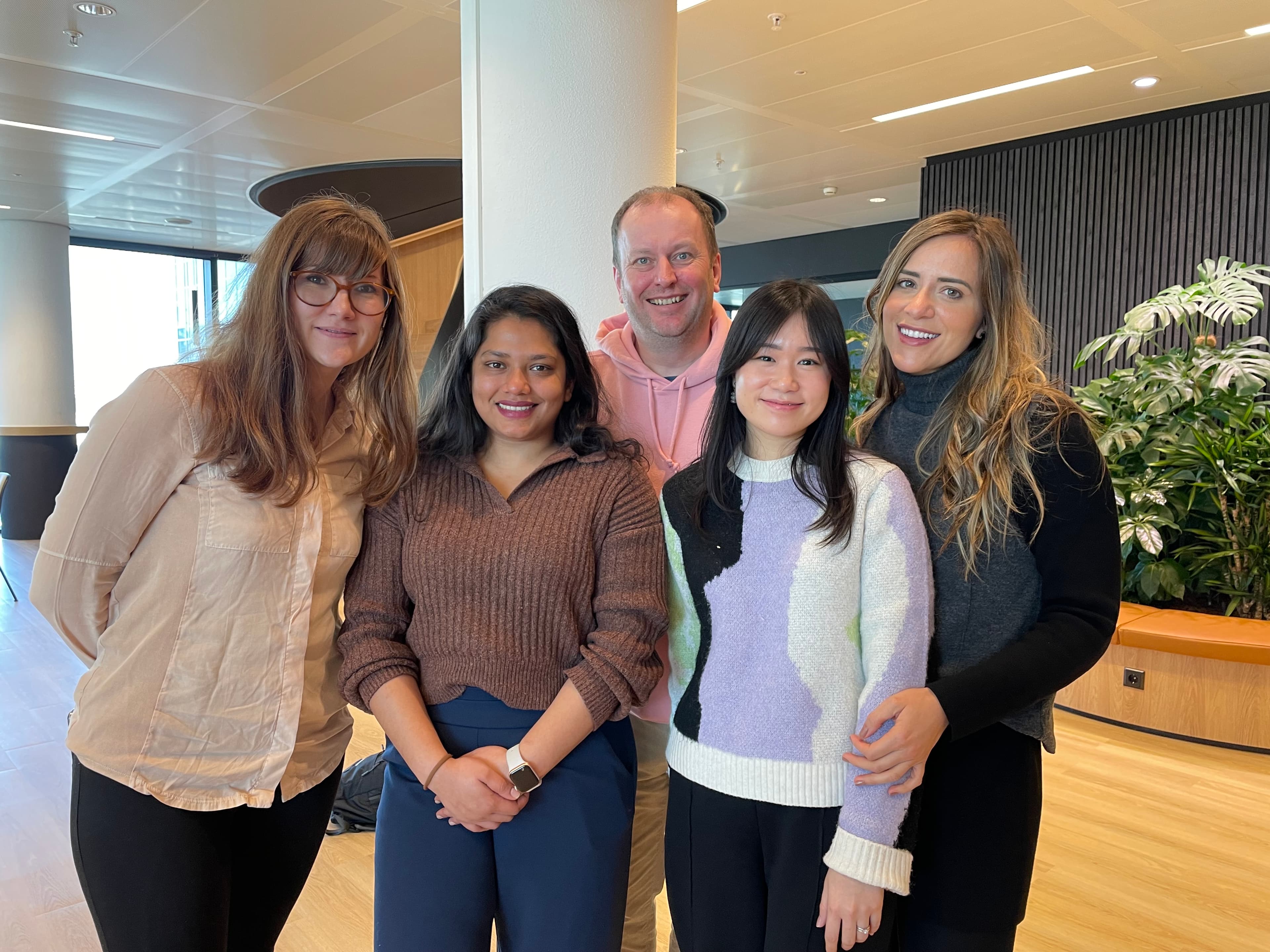 A group of five people standing indoors, smiling at the camera. They are in a modern office space with plants and large windows.
