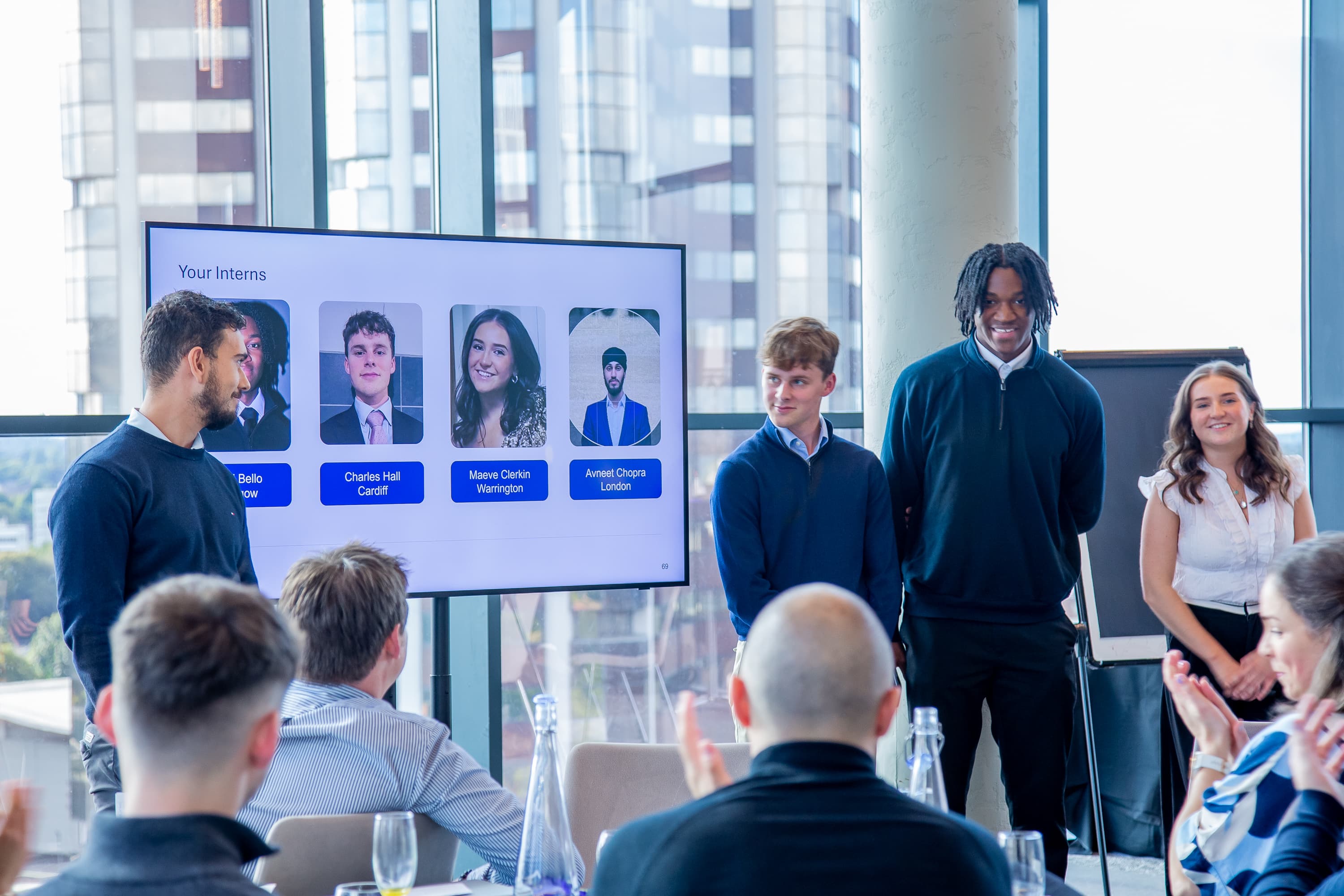 Four people present to an audience in a modern office space. A screen displays intern profiles and their names.