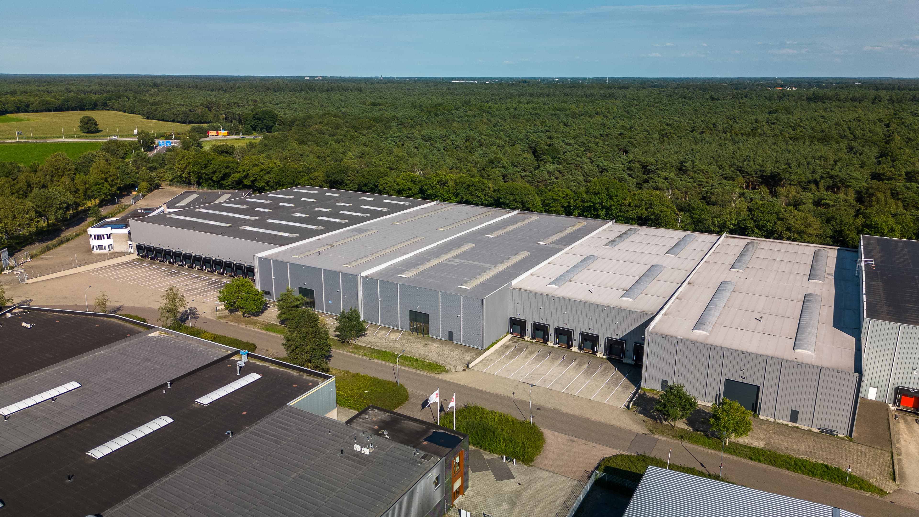 Aerial view of a large industrial warehouse complex surrounded by trees, with loading bays and adjacent buildings under a clear sky.