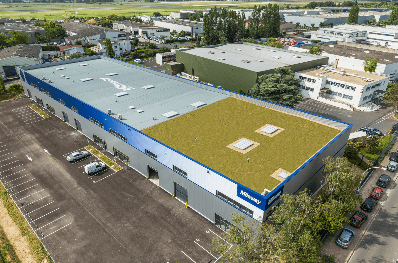 Aerial view of a large industrial building with a green roof, surrounded by parking and other industrial structures.