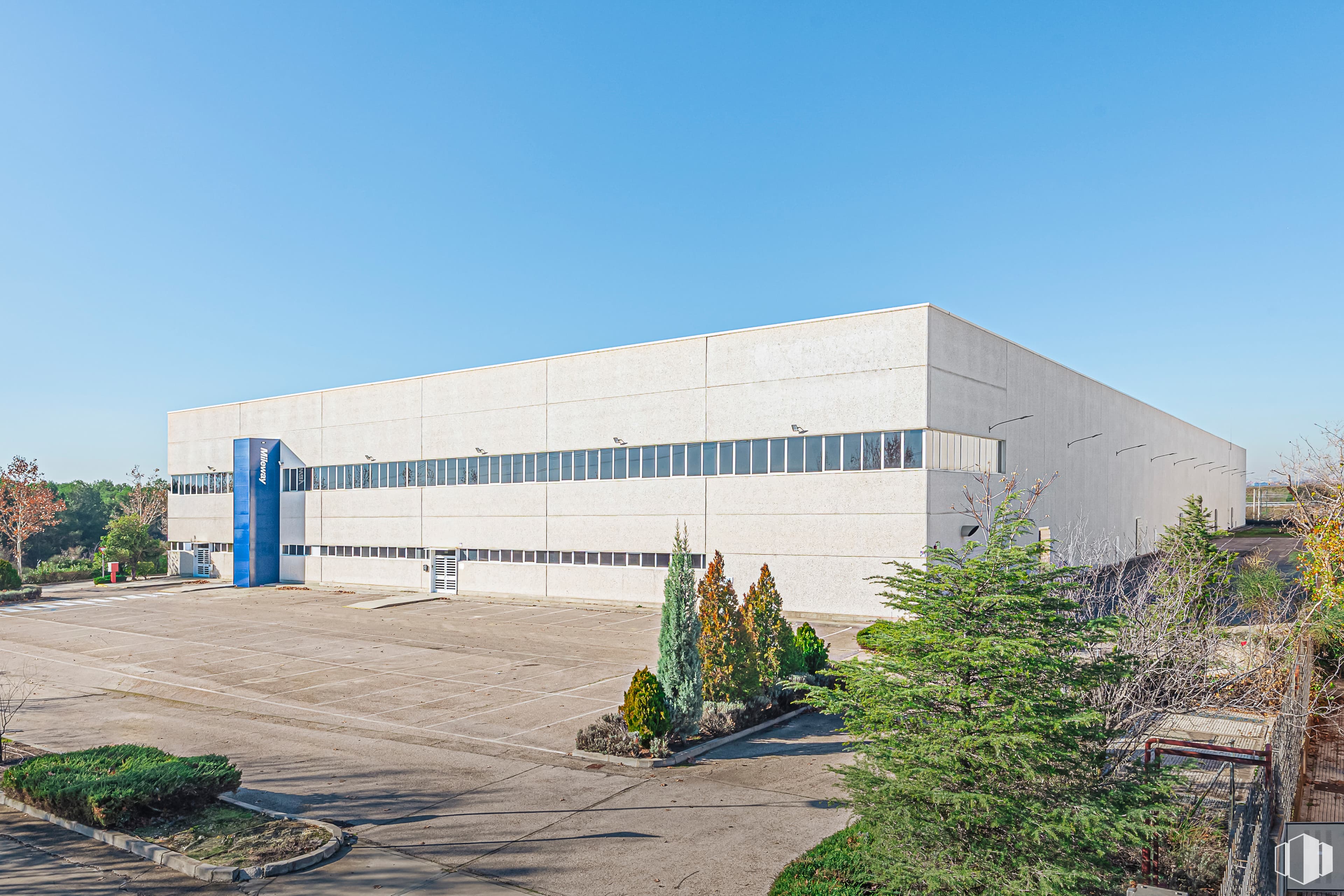 A large, white industrial warehouse with blue accents stands under a clear blue sky, surrounded by sparse greenery and a paved parking area.