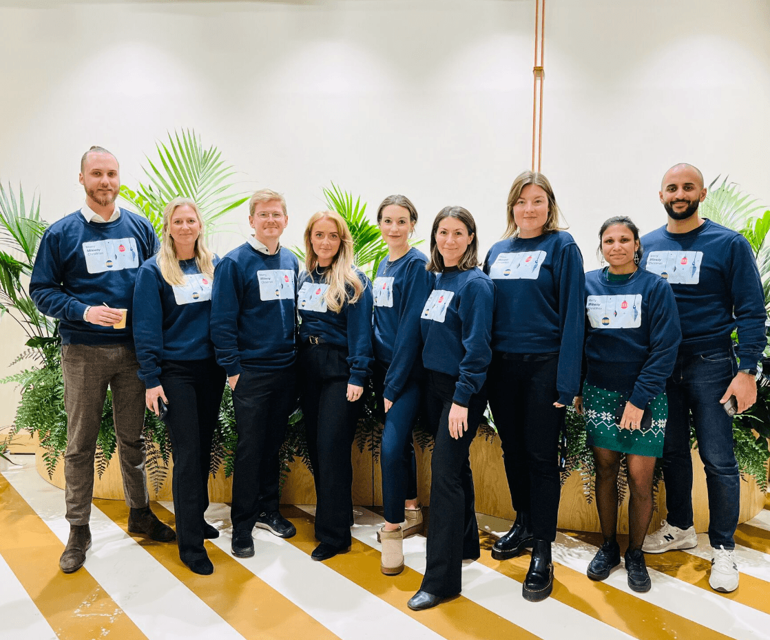 A group of nine people standing indoors, wearing matching navy sweatshirts. There are potted plants behind them.