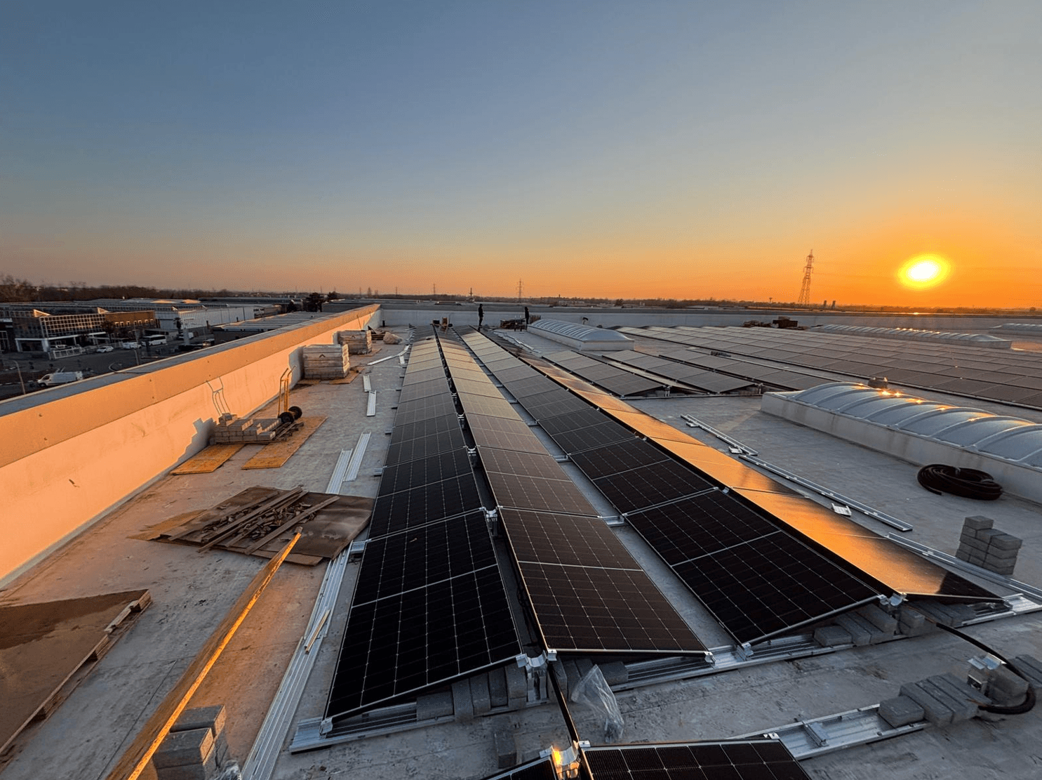 Rooftop solar panels during sunset, reflecting sunlight. Urban industrial background with a clear sky and power lines on the horizon.