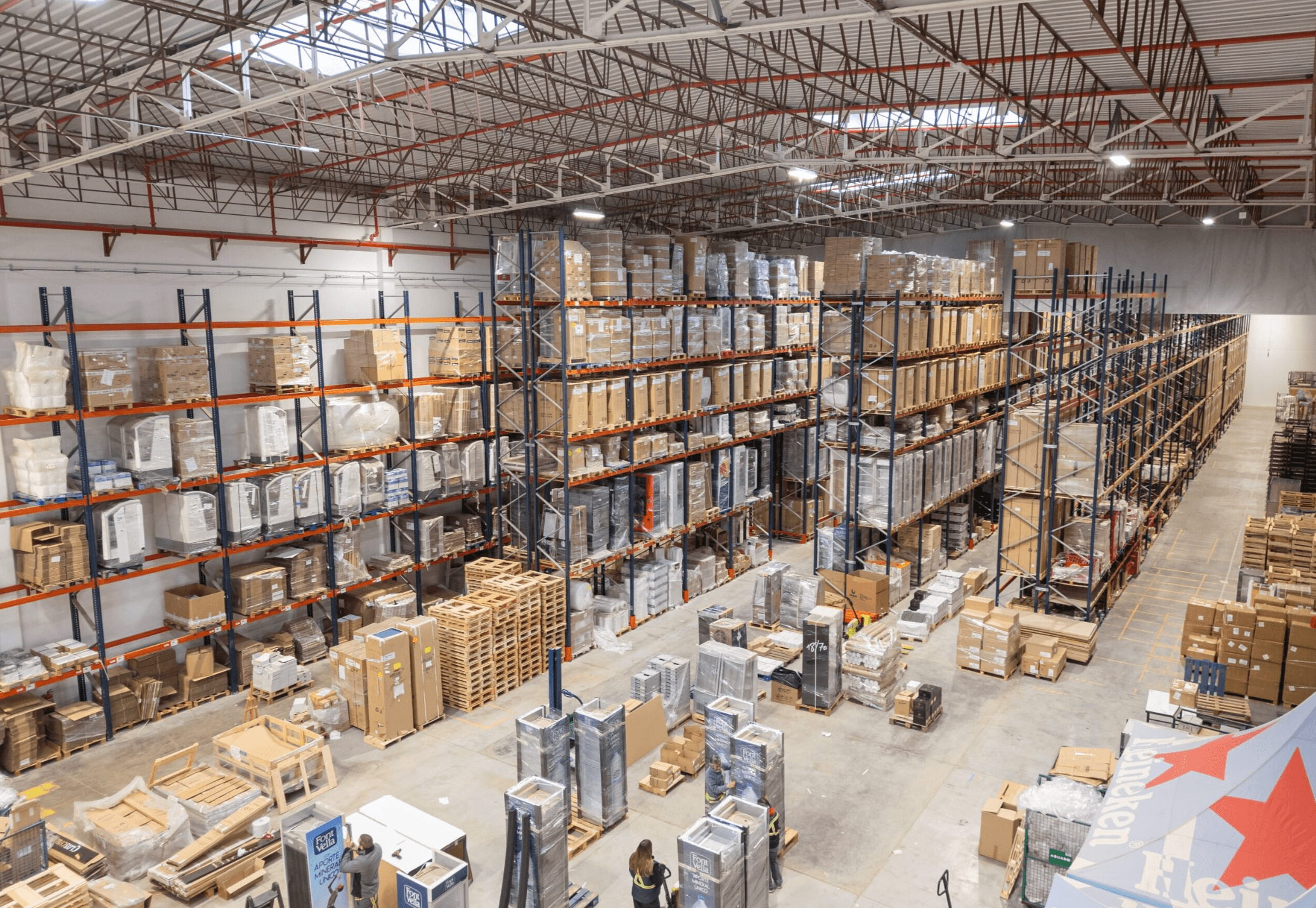 Interior of a large warehouse with high shelves stocked with various boxes and items. Forklifts and workers are visible on the floor, organizing and moving goods.