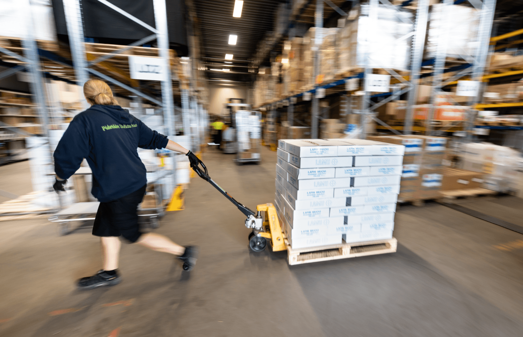 Person pulling a pallet jack loaded with boxes through a warehouse, wearing a dark jacket and shorts.
