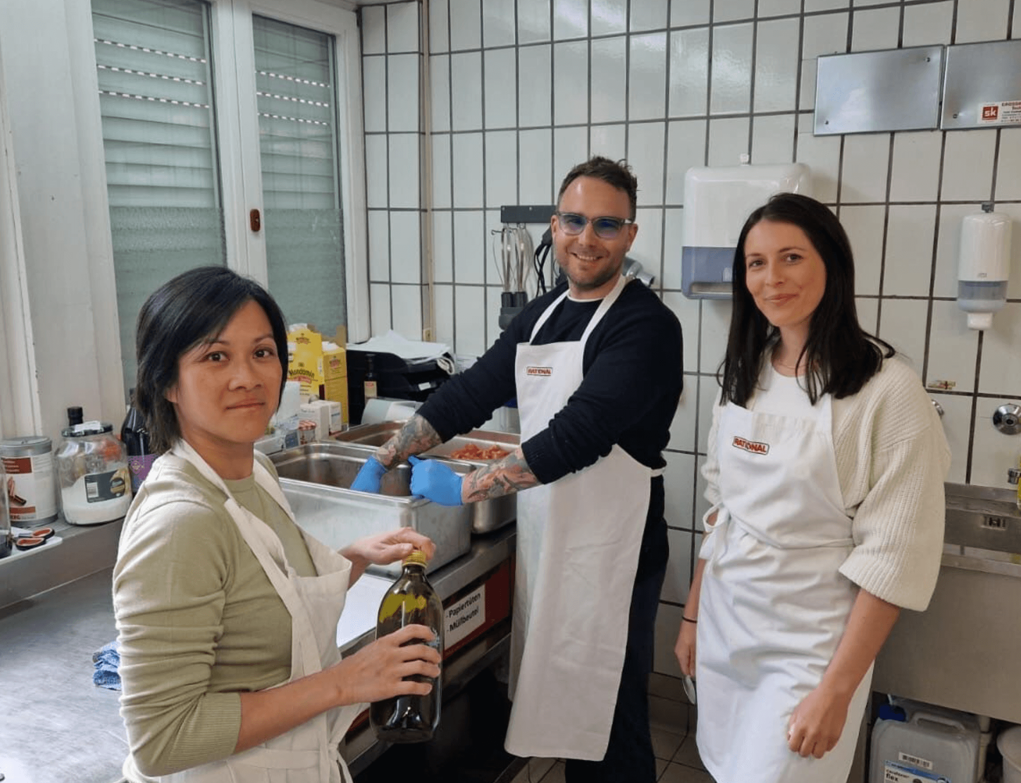 Three people wearing aprons are in a kitchen. One holds a bottle, another is washing dishes, and the third stands nearby.