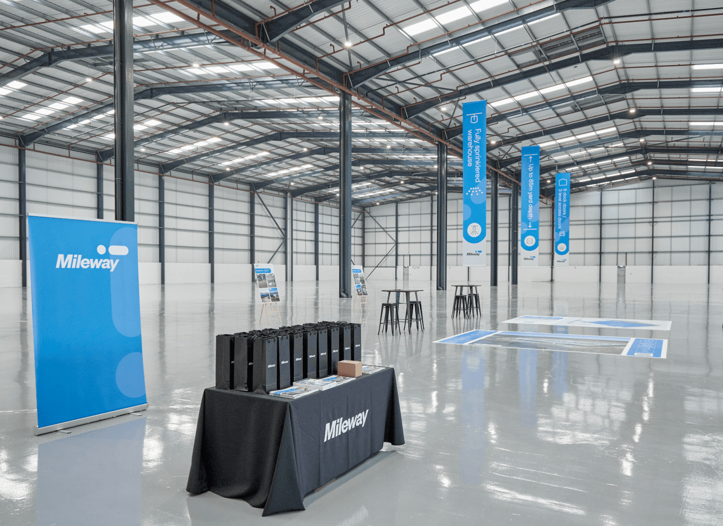 Large, empty warehouse with high ceilings and skylights. A Mileway-branded table with bags and brochures stands near banners and a few high tables and stools.