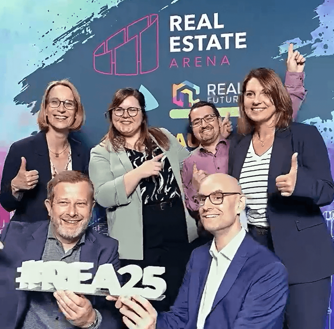 Six people pose and smile at the Real Estate Arena event, with two holding a #REA25 sign in front of a branded backdrop.