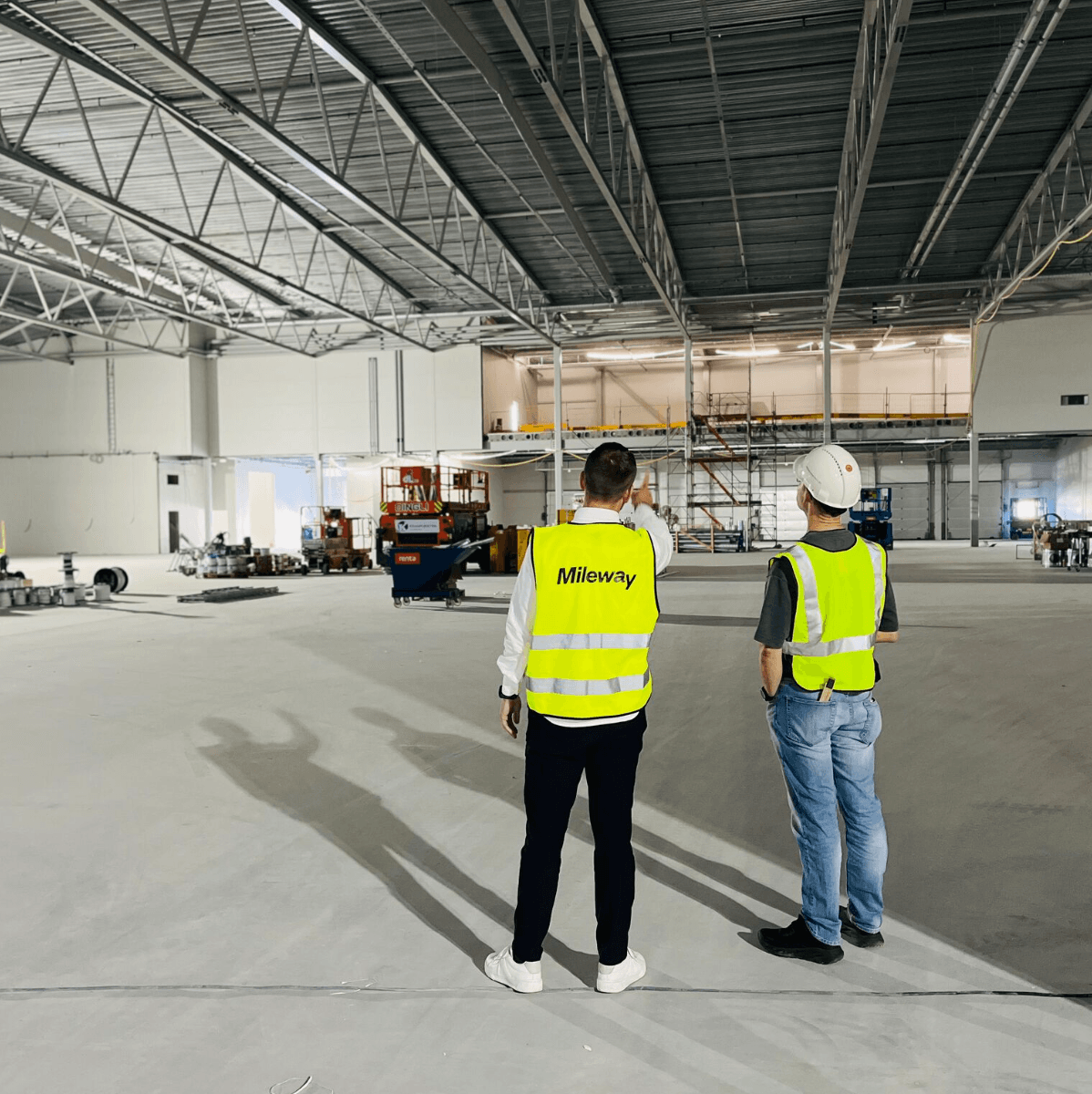 Two workers in high-visibility vests and helmets stand inside a large, unfinished industrial building, observing construction progress.