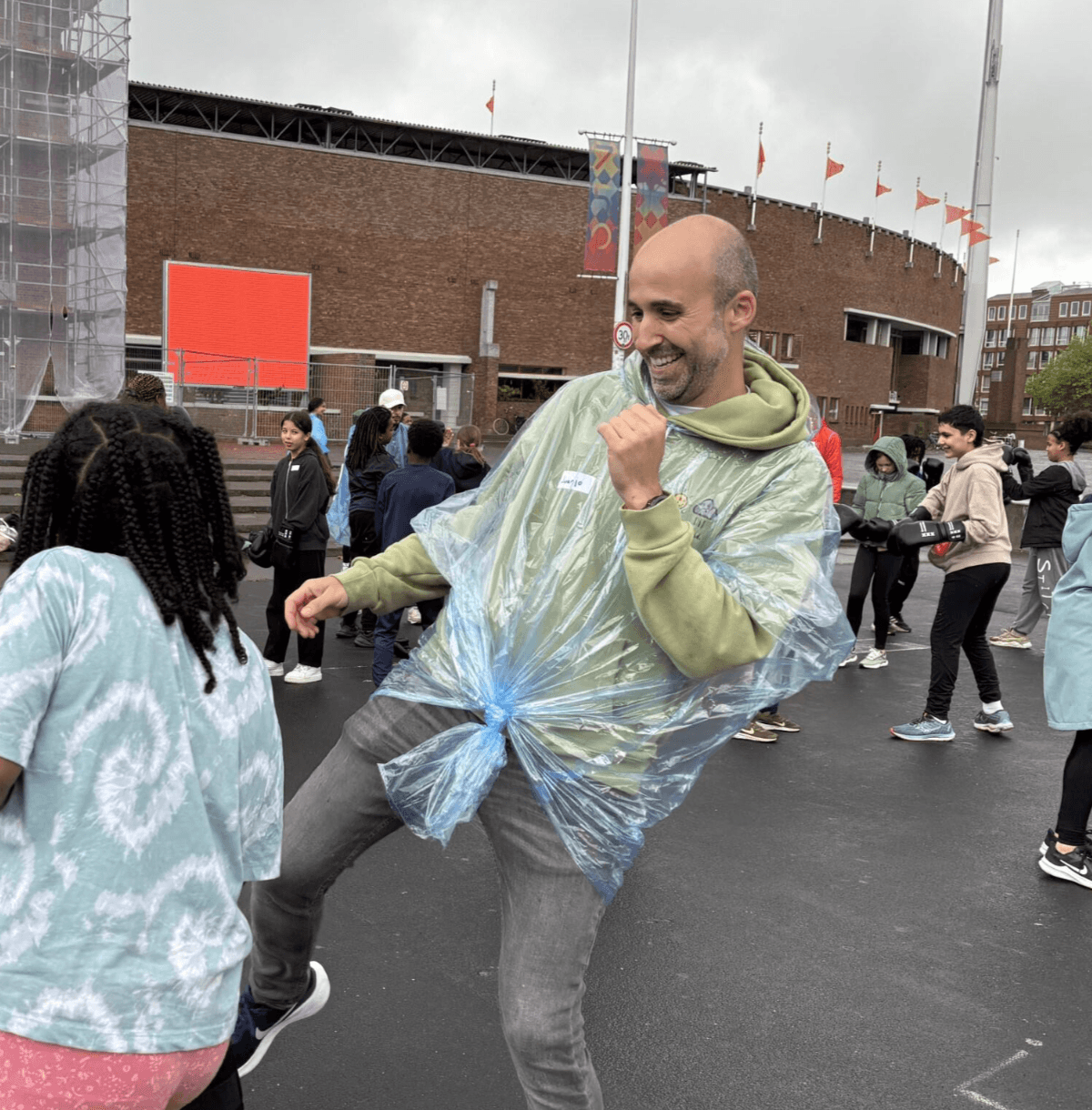 A man wearing a plastic poncho and jeans dances outdoors with others on a cloudy day. People in casual clothes are in the background near a brick building.