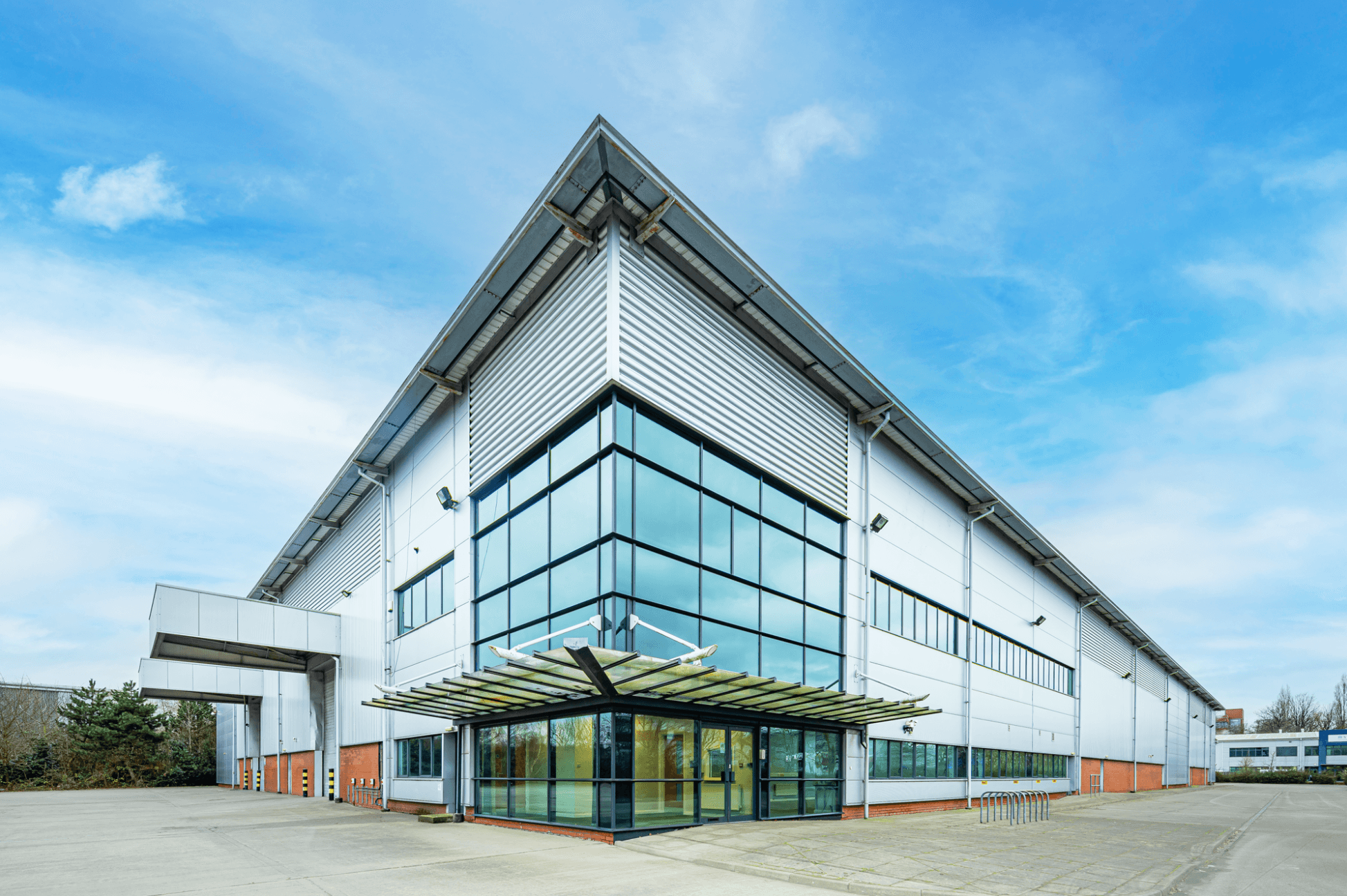 Modern industrial warehouse with large glass windows and metal siding under a blue sky, featuring a covered entrance and empty surrounding pavement.