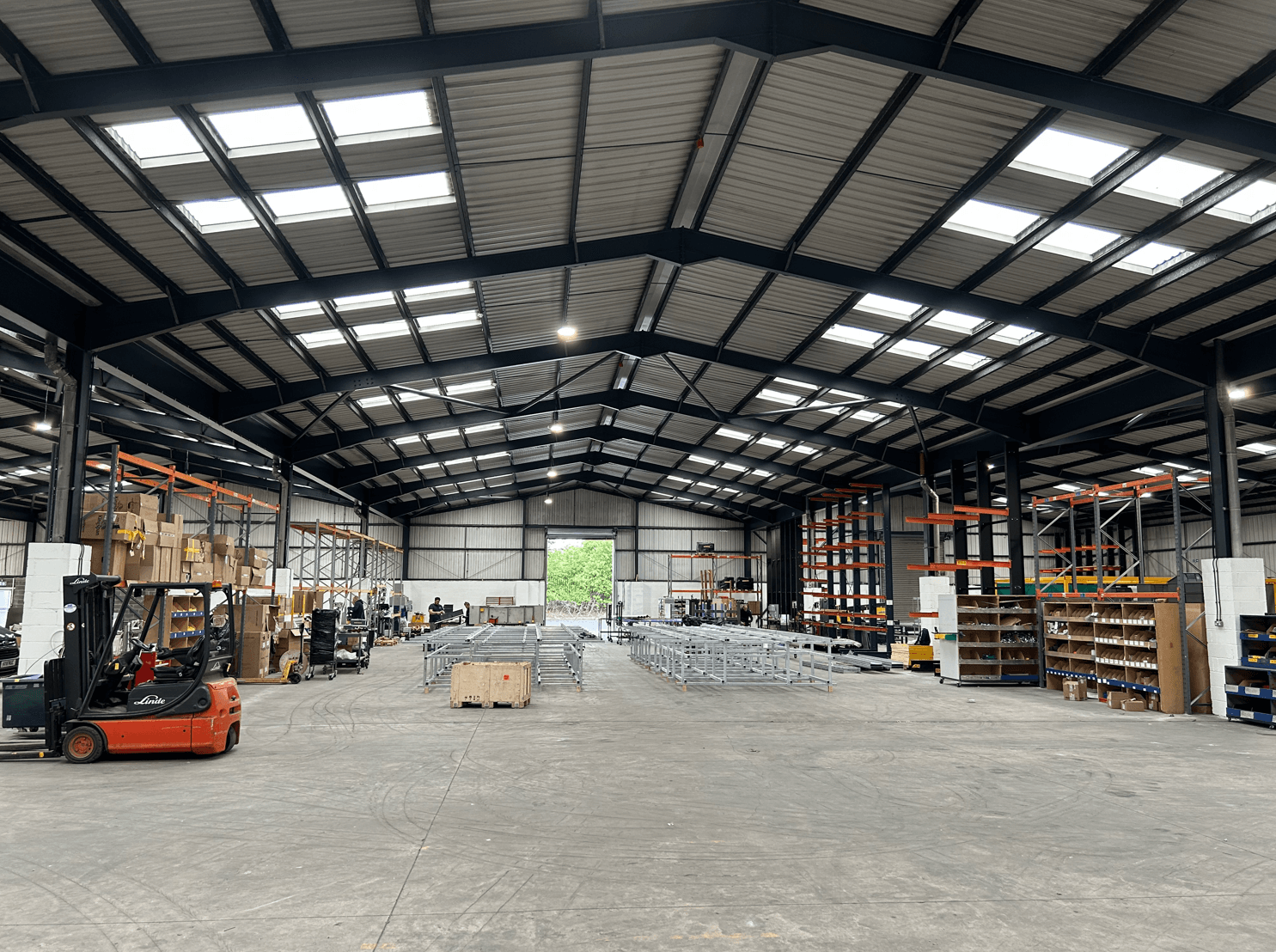 Large warehouse interior with high ceilings, shelving units, pallets, metal structures, and forklifts; workers are operating machinery and organizing materials.
