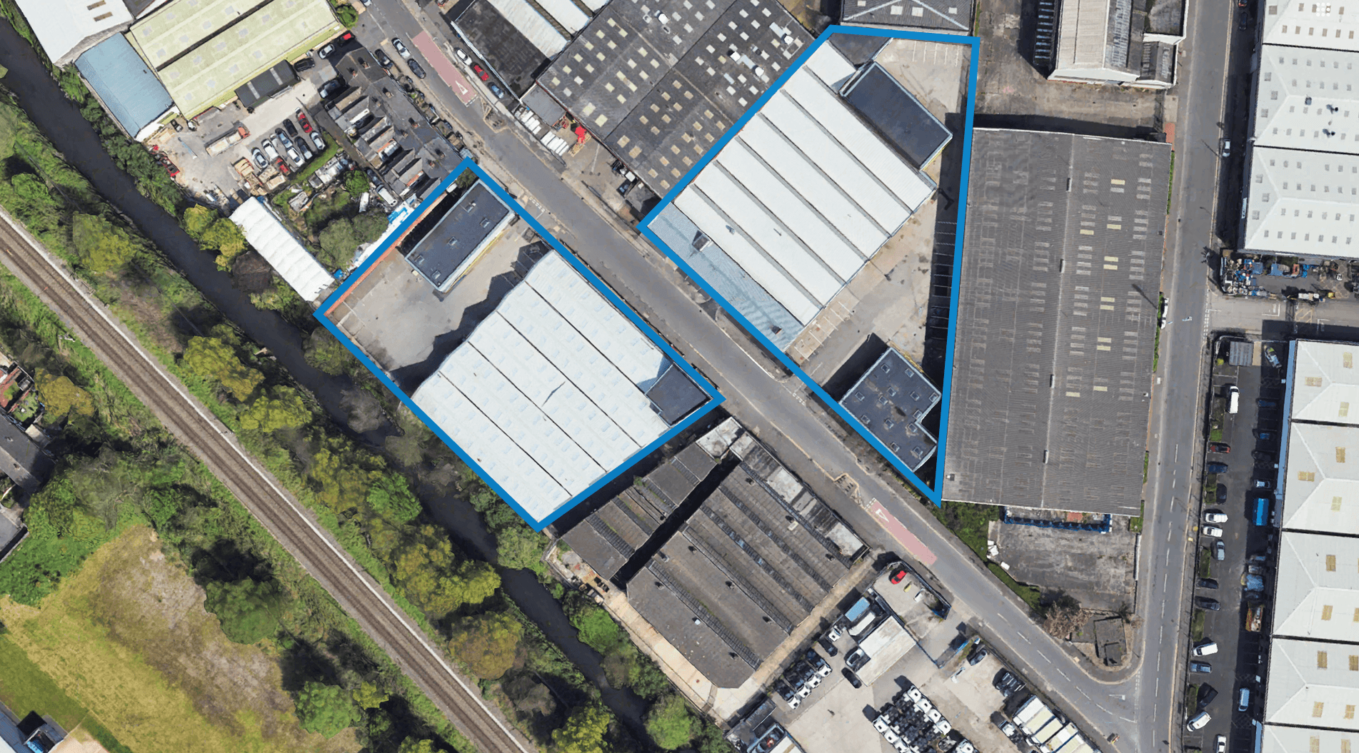 Aerial view of an industrial area with multiple large warehouses and factories; two adjacent sites are outlined in blue along a road near a canal and railway line.