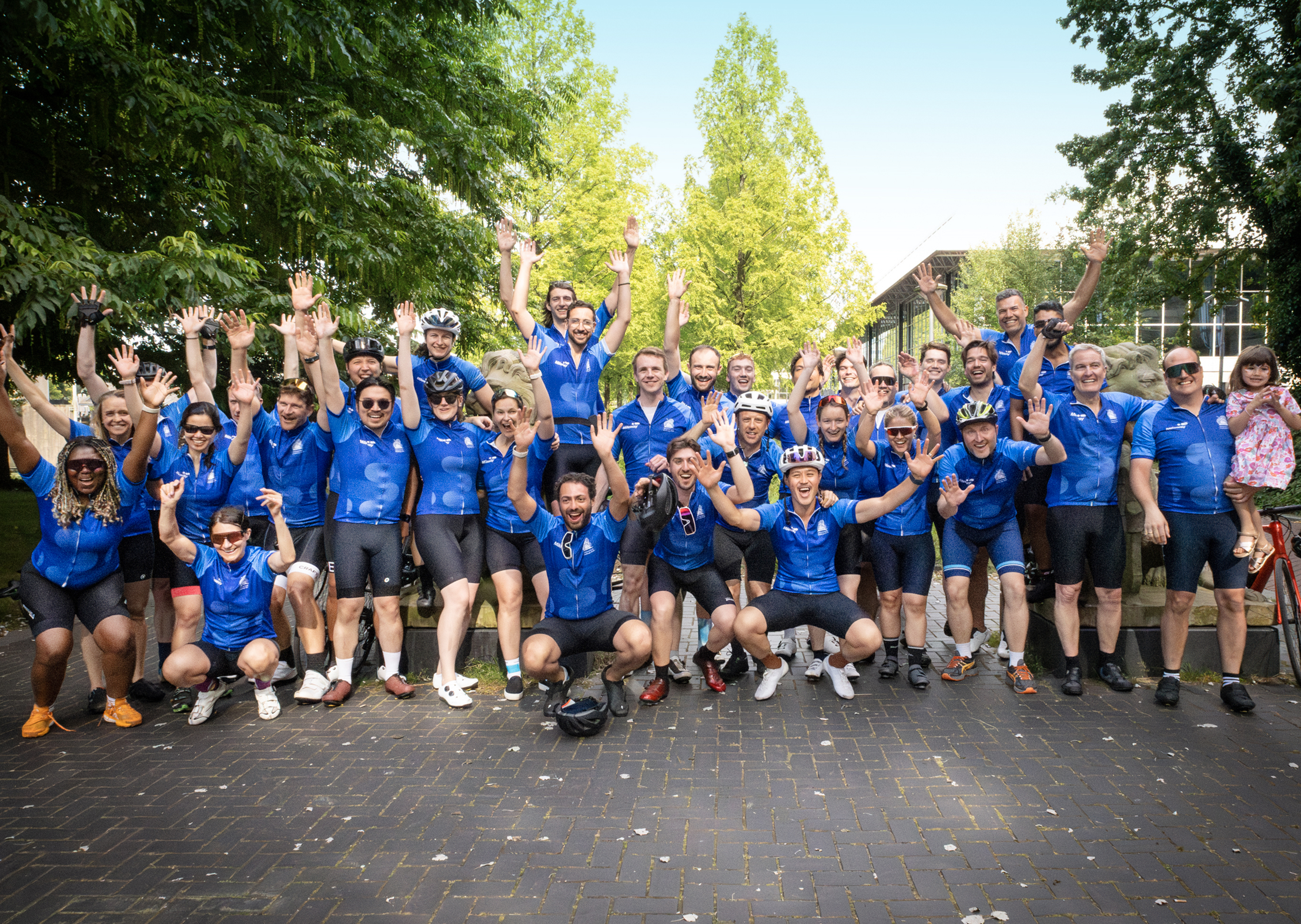 A large group of cyclists in matching blue jerseys pose outdoors with raised arms, smiling for a group photo on a paved path surrounded by greenery.