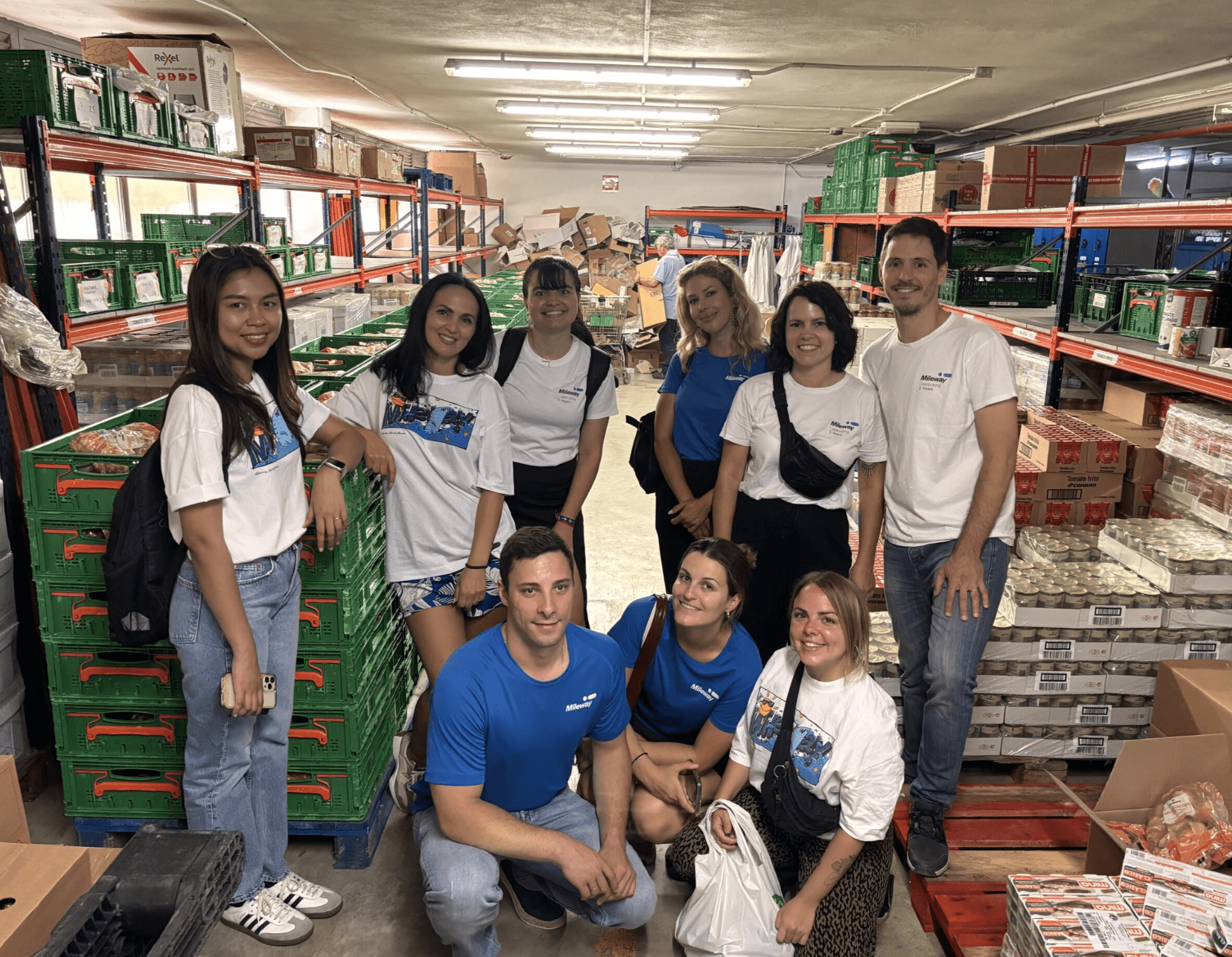 A group of people wearing white and blue shirts pose together inside a warehouse lined with shelves of food products and green crates.