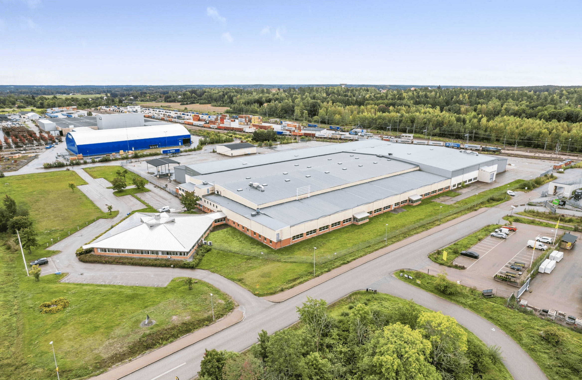 Aerial view of a large industrial building with adjacent office, parking lot, and surrounding greenery; other industrial facilities visible in the background.