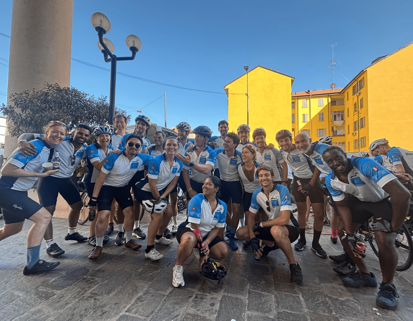 A group of cyclists wearing matching jerseys pose and smile together on a cobblestone street with yellow buildings in the background.