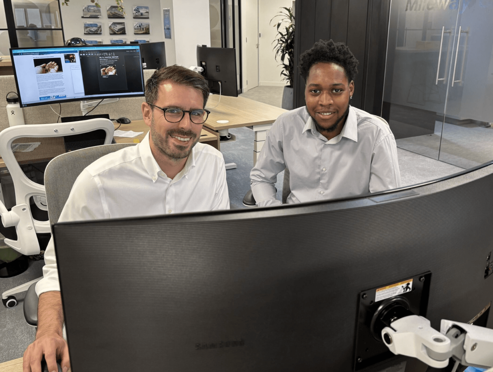 Two men in white shirts are seated at a desk, smiling at the camera, with a large computer monitor and office workspace in the background.