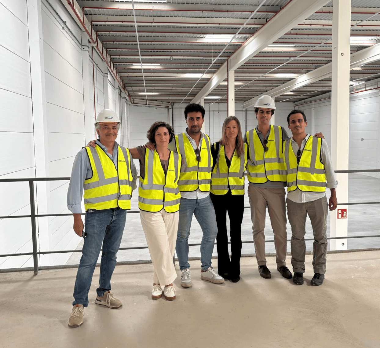 Six people wearing safety vests and hard hats stand together inside a large, unfinished industrial building.