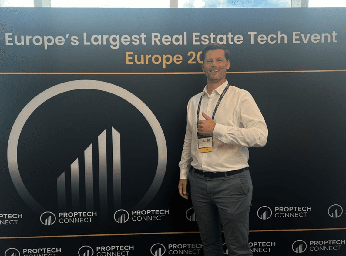 A man in business attire stands smiling with a thumbs up in front of a Europes Largest Real Estate Tech Event PropTech Connect banner.