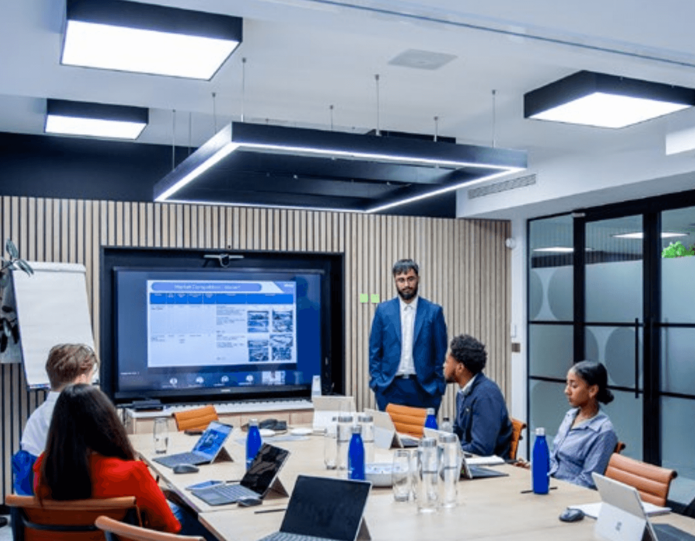 A group of people in a modern conference room attend a presentation; a man stands by a large screen displaying charts and images whilst others sit at a table with laptops.